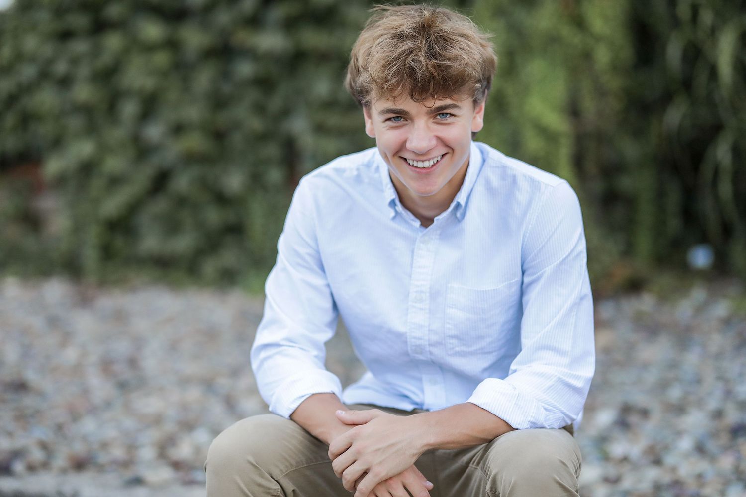 Someone in a light blue button-down shirt and khaki pants sits outdoors smiling against a natural background.