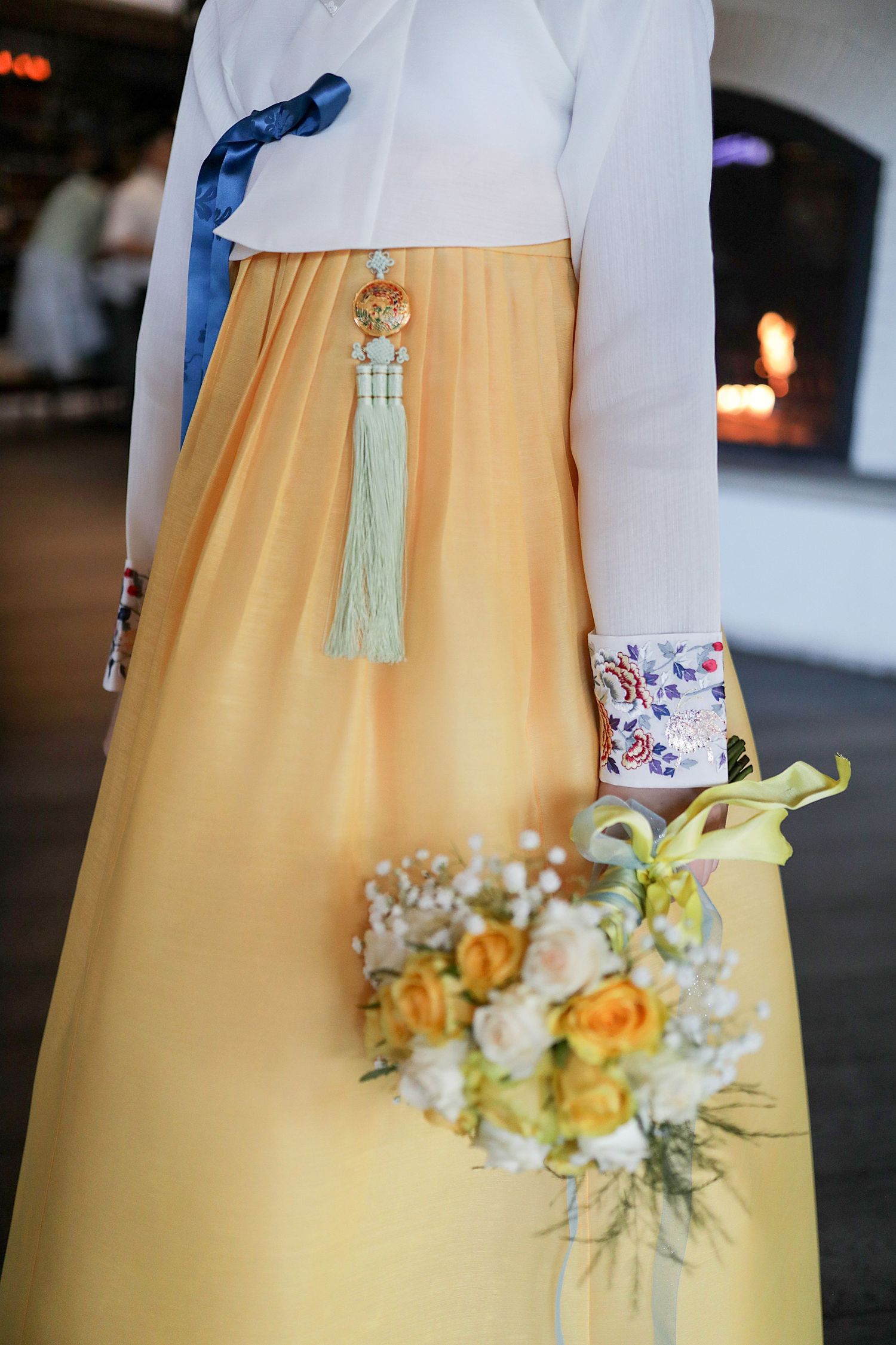 A yellow traditional Korean hanbok dress adorned with a mint tassel and blue accent, with a floral bouquet of yellow and white.