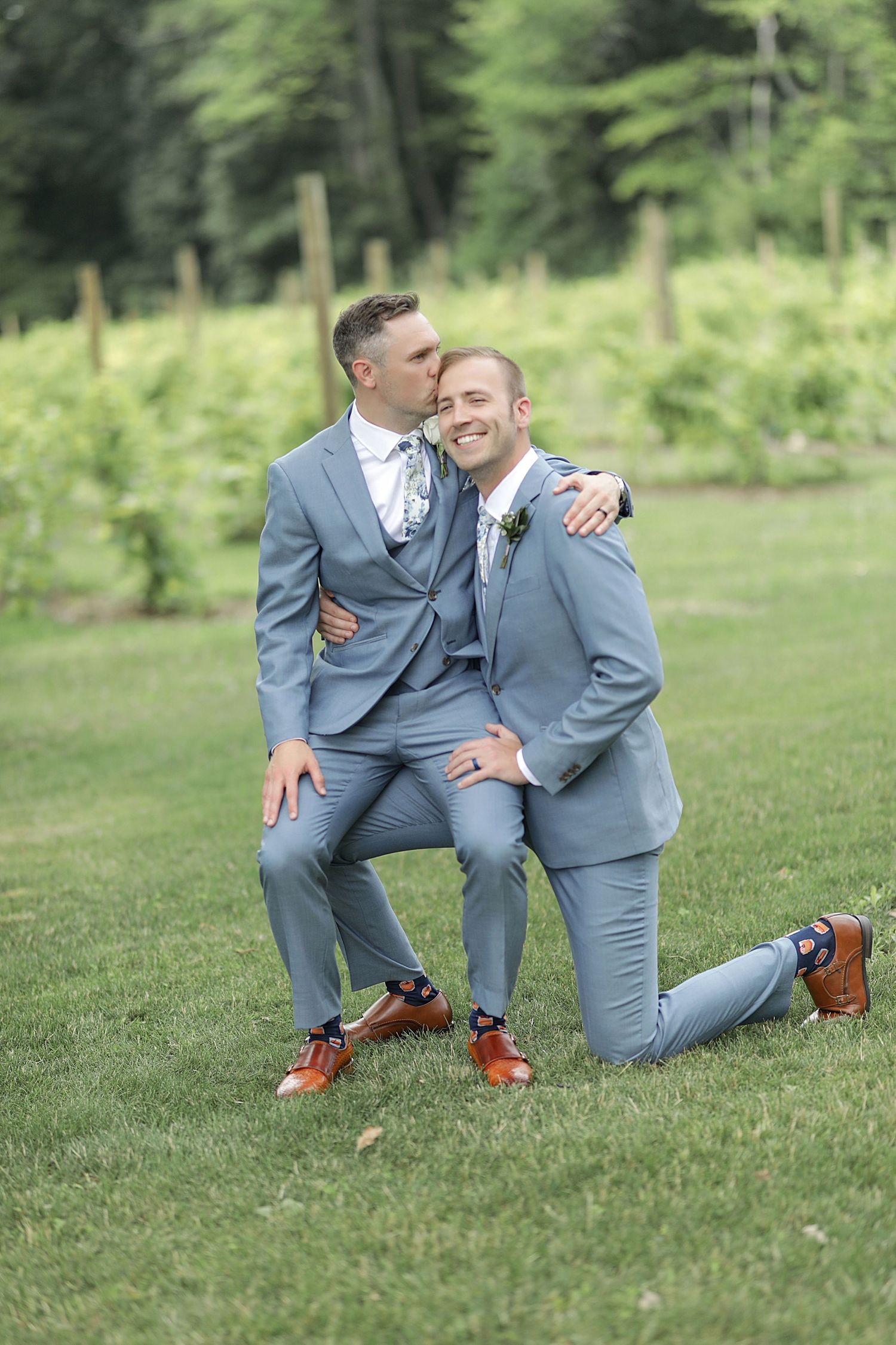 A playful wedding photo of two people in matching gray suits on a grassy lawn.