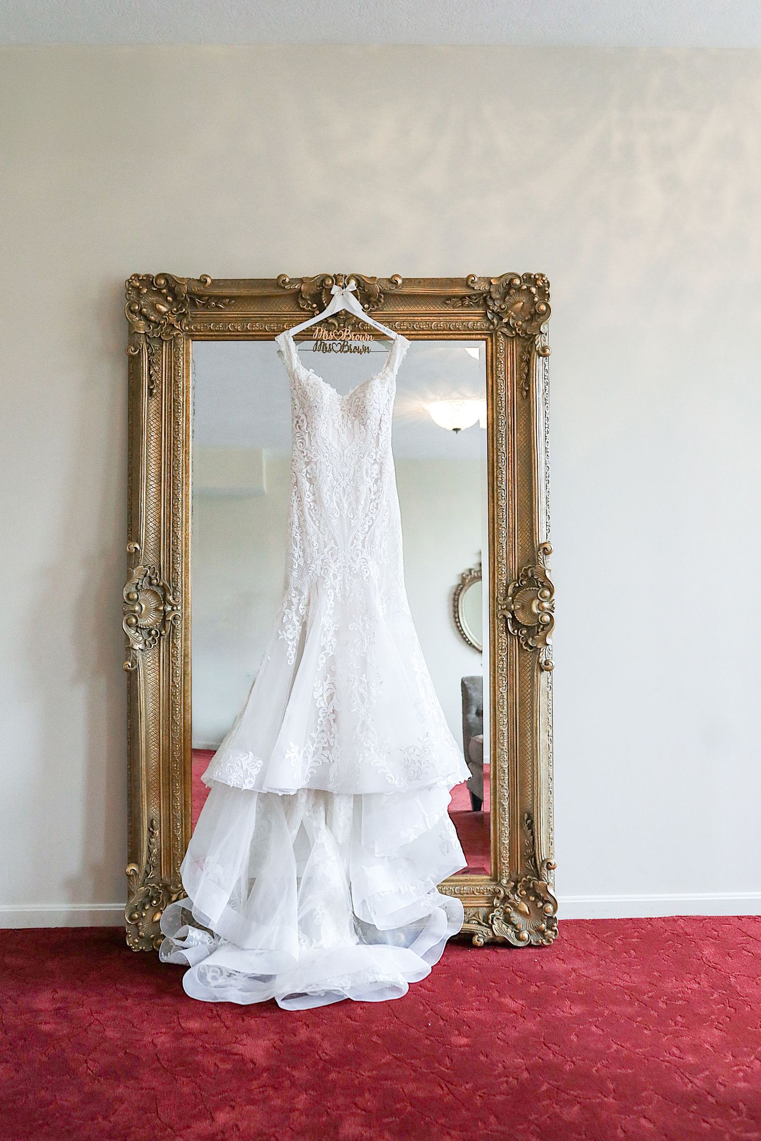 White lace wedding dress displayed on a hanger against an ornate gold-framed mirror with red carpet below.