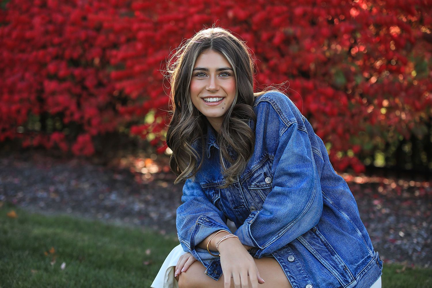 A smiling person wearing a denim jacket poses outdoors with vibrant red autumn foliage in the background.