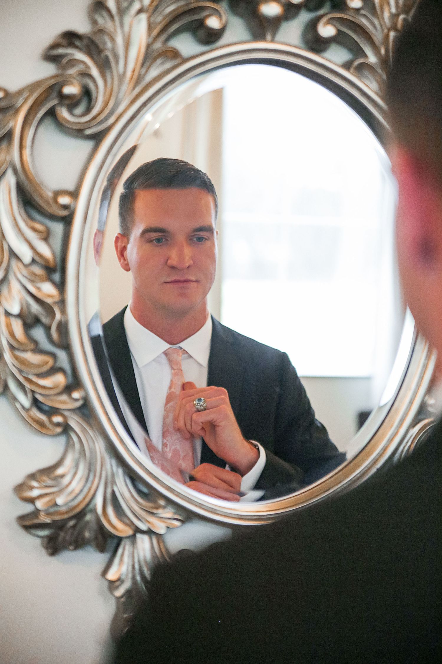 A reflection in an ornate silver mirror shows someone in formal attire adjusting their tie.