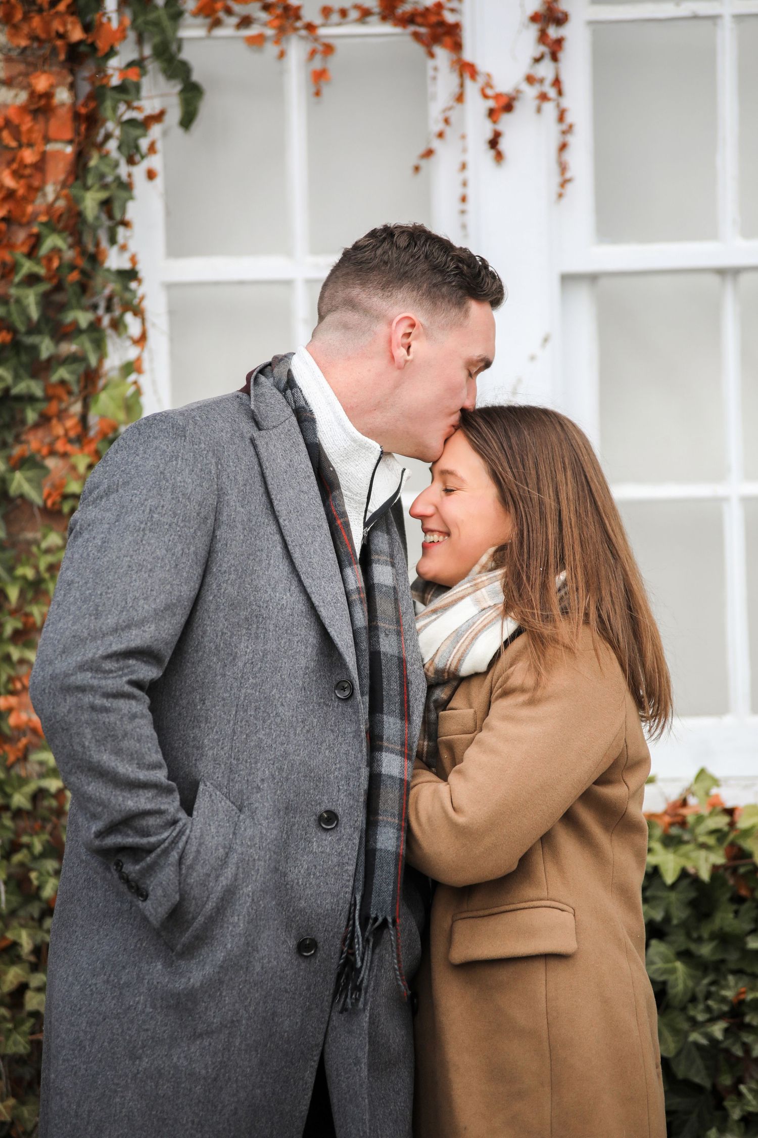 A romantic couple in winter coats share a tender moment against a white building with autumn ivy.