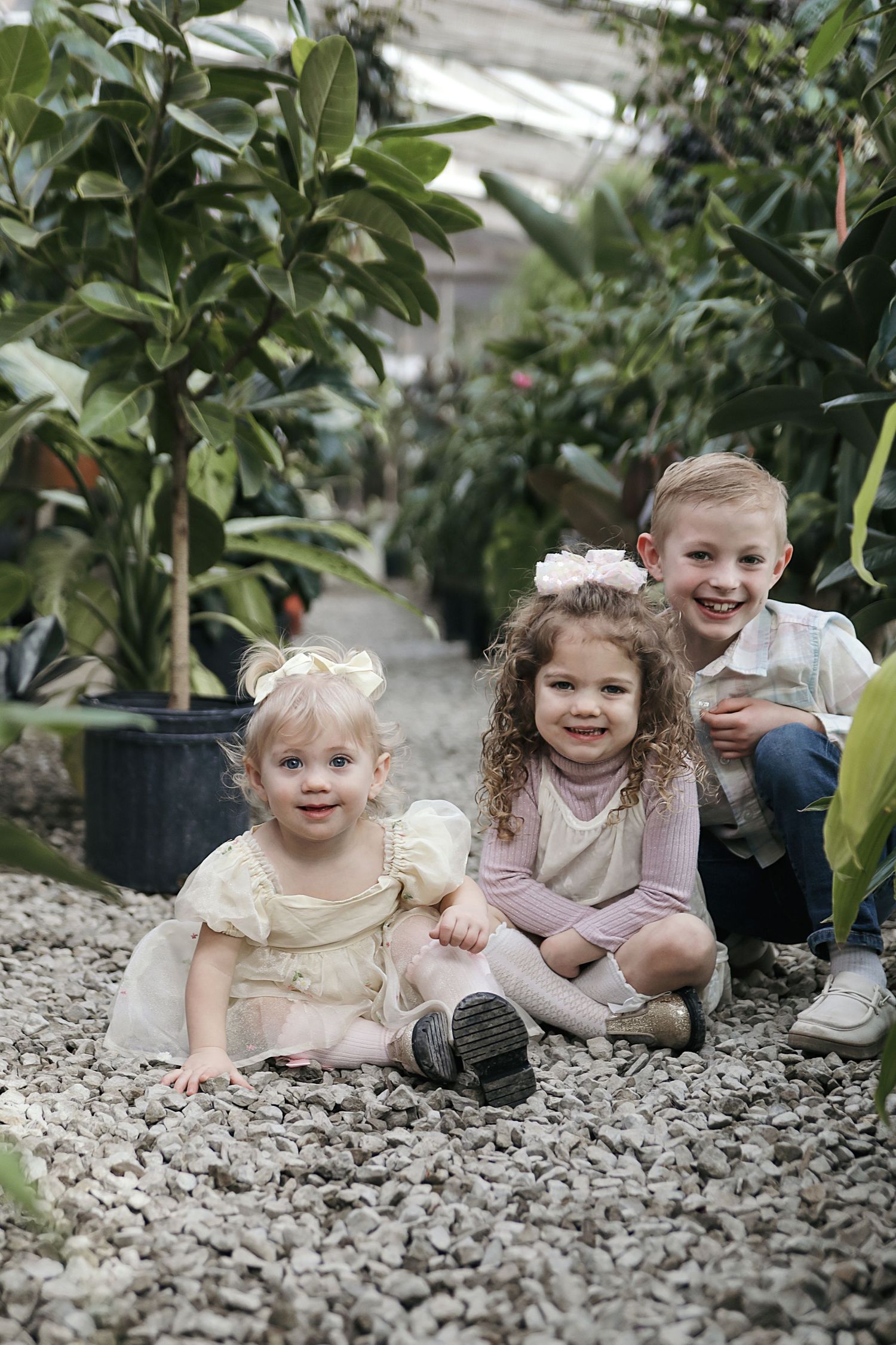 Three young children sitting together on gravel path in a greenhouse surrounded by potted plants.
