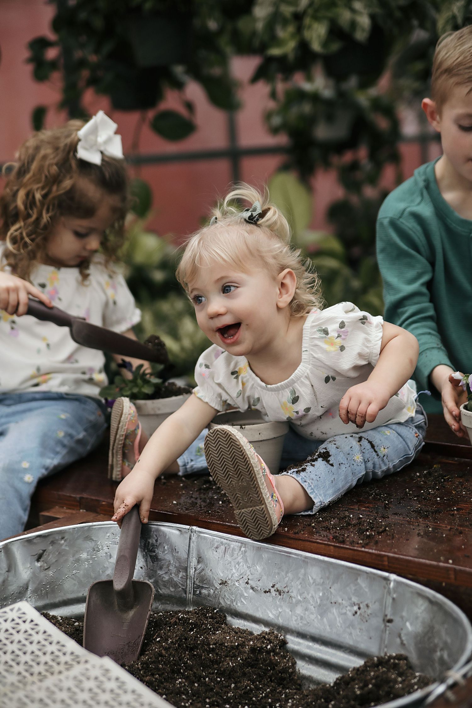 A small child in a white shirt plays with soil and gardening tools in an outdoor planter box.