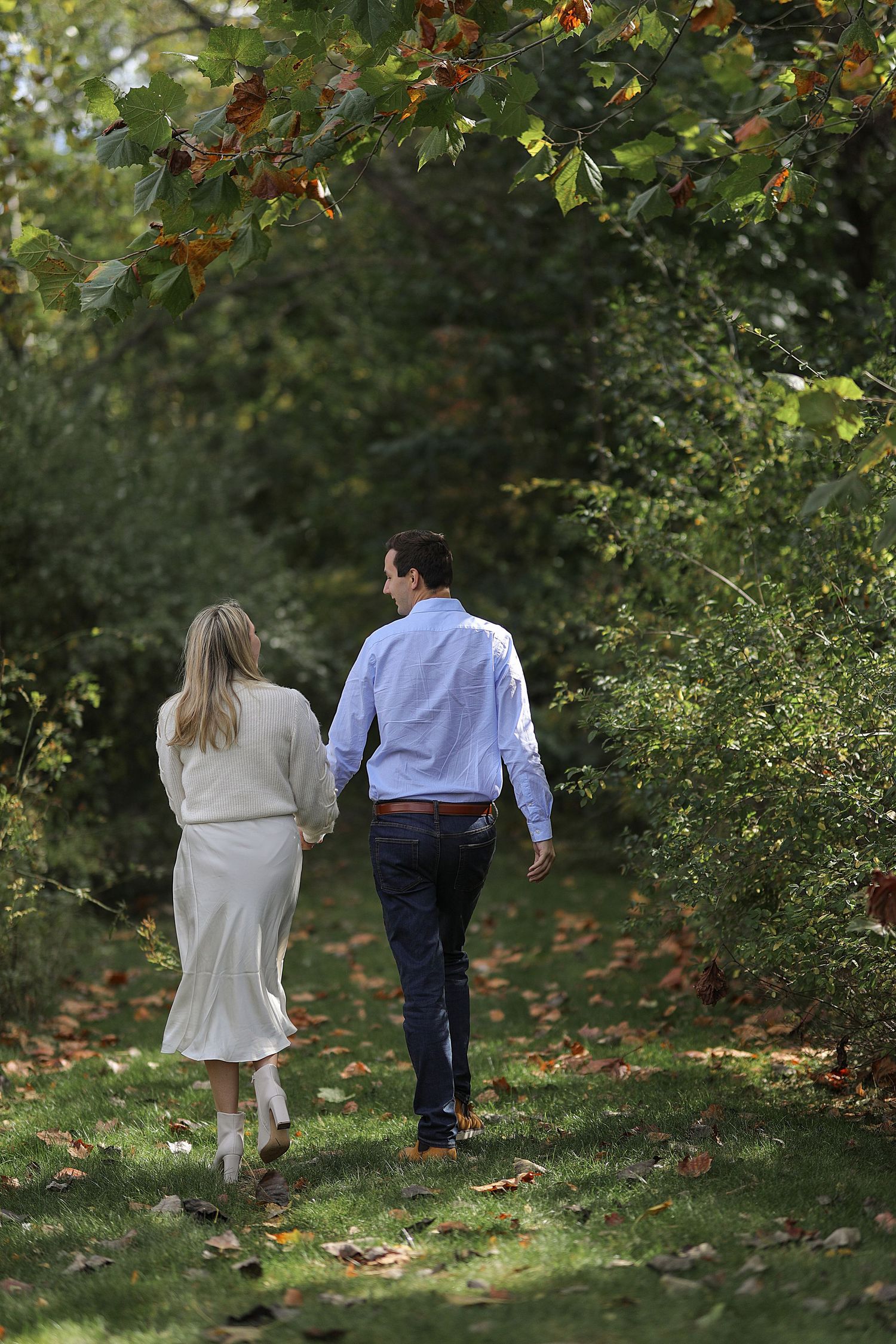 A couple walks hand in hand down a leaf-strewn garden path beneath autumn trees and foliage.