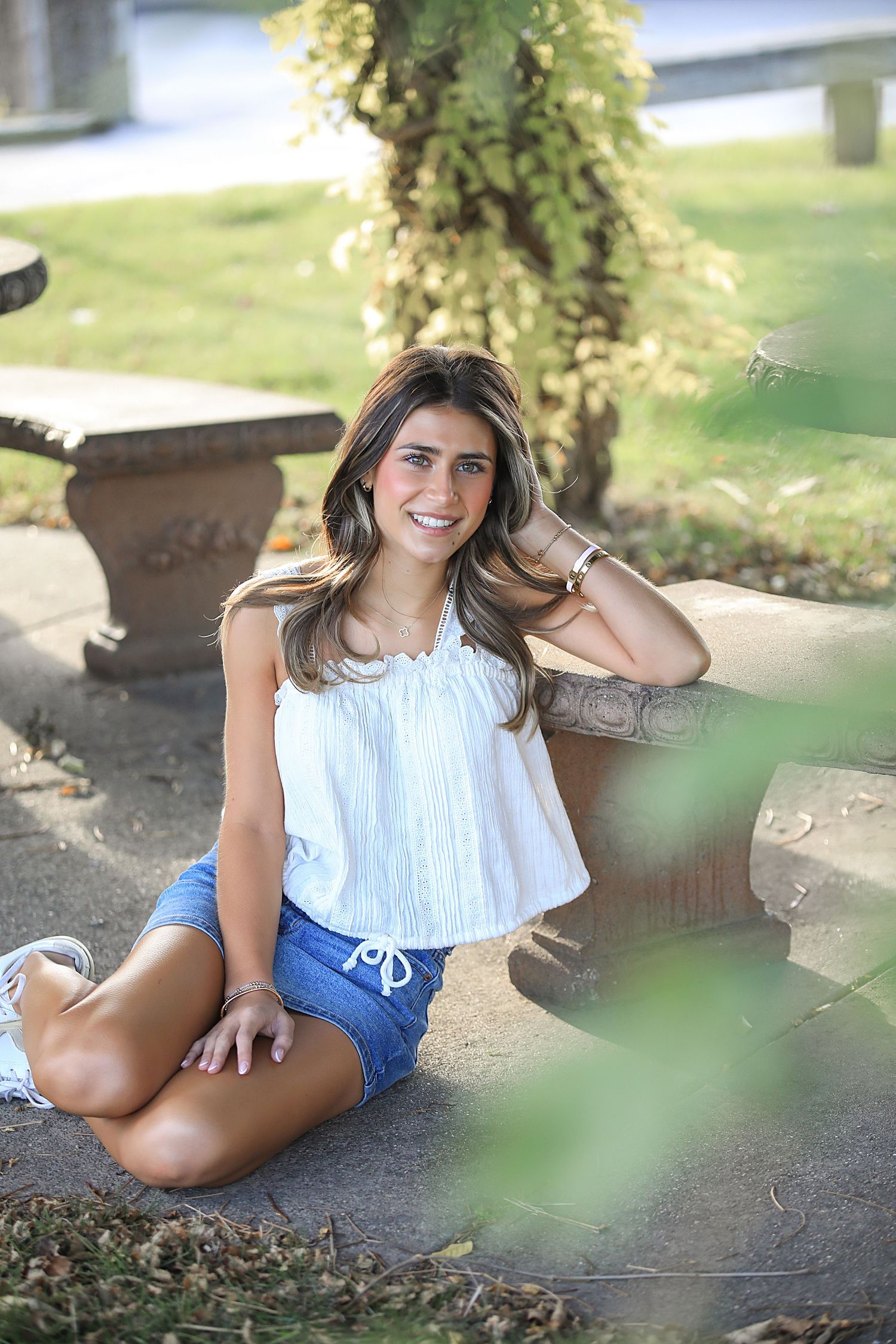 A person in a white top and denim shorts sits casually outdoors on concrete steps near garden planters and greenery.