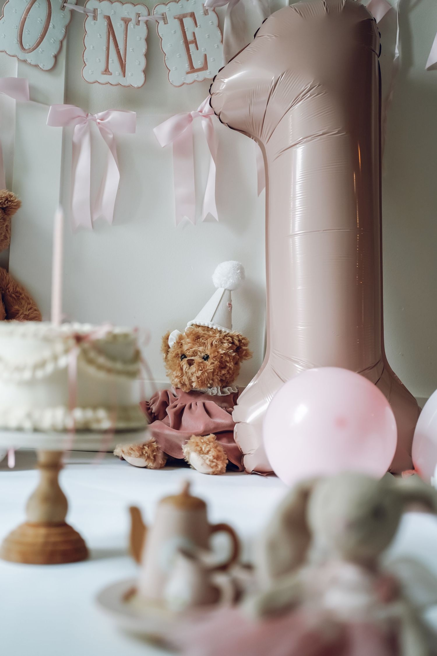 First birthday party decor with large pink number one balloon, teddy bear, and vintage tea cups set on white table.