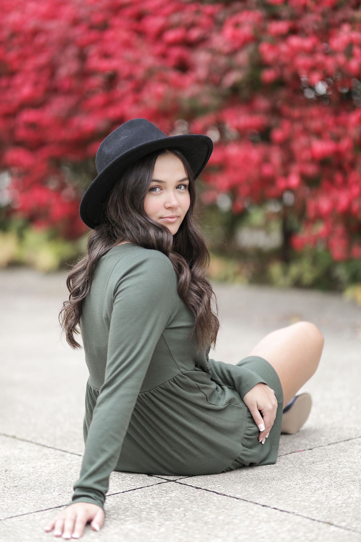 Woman in green dress and black hat poses outdoors against vibrant red autumn foliage.