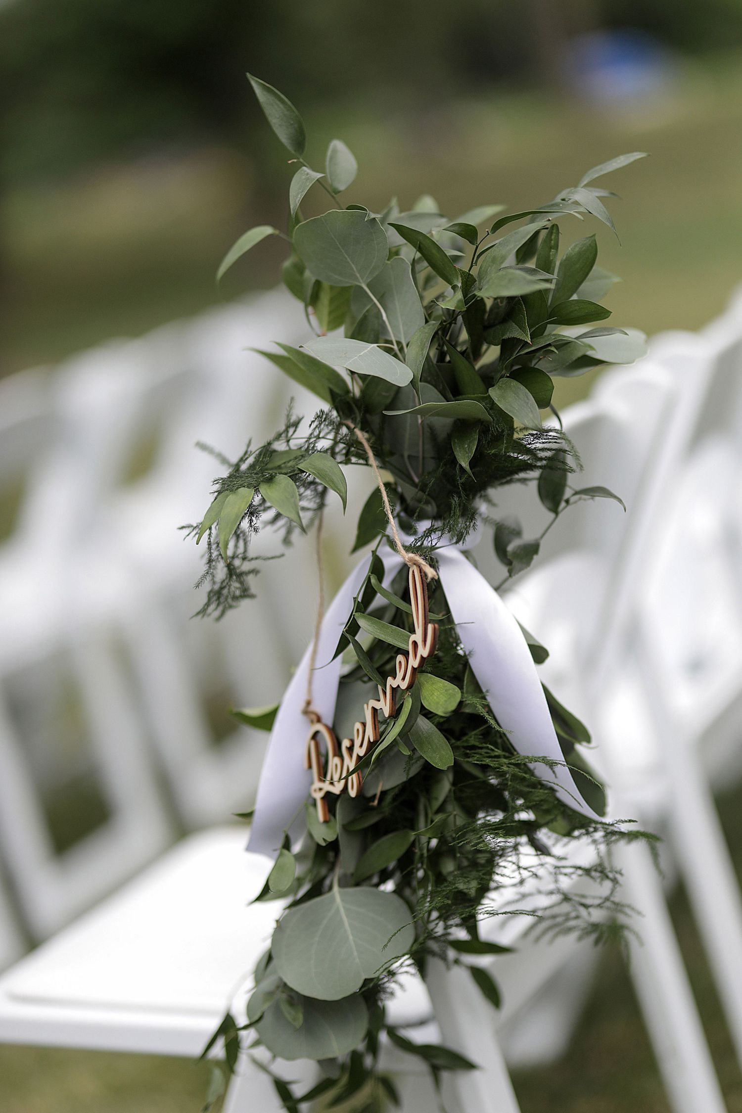 White folding chairs decorated with greenery and ribbons line up for an outdoor wedding ceremony.