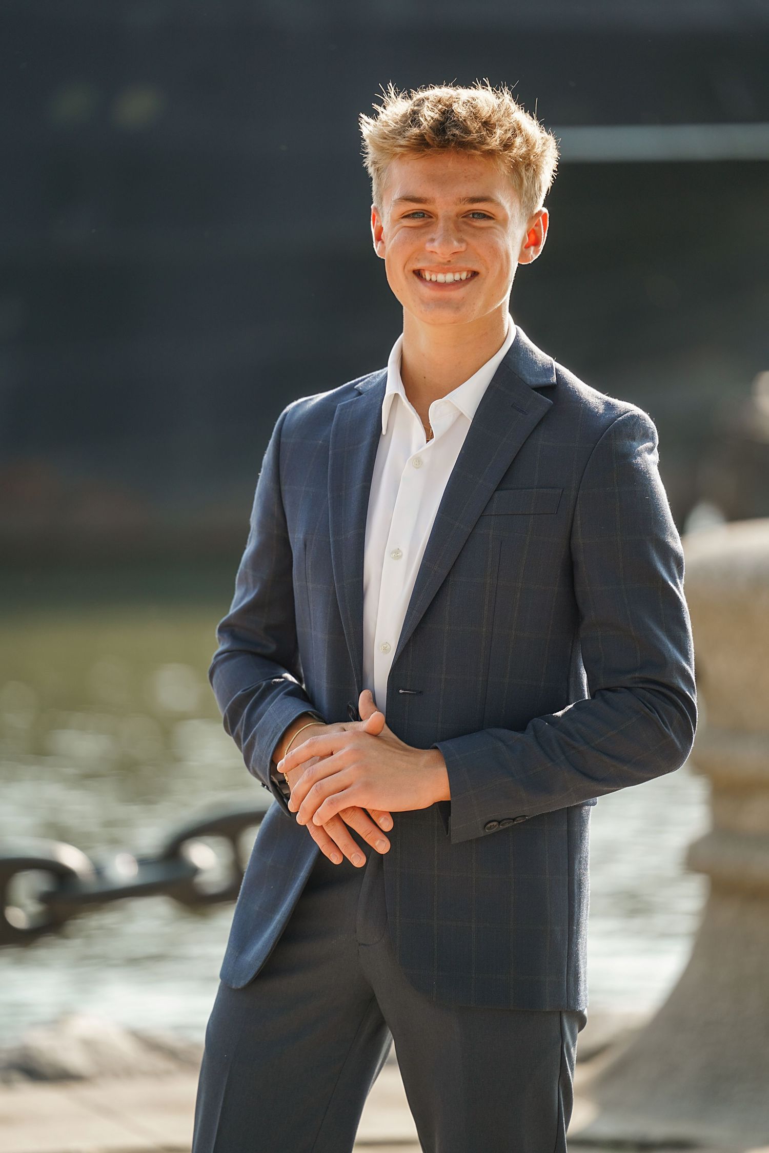Young person wearing a navy suit jacket and white shirt poses casually outdoors by a waterfront in multiple shots.