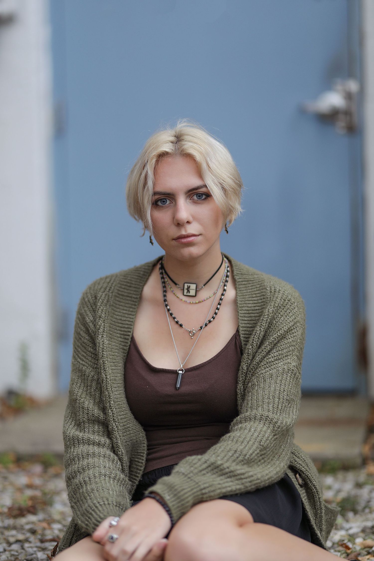 Portrait of someone in an olive green cardigan over brown top with layered necklaces against a blue wall background.
