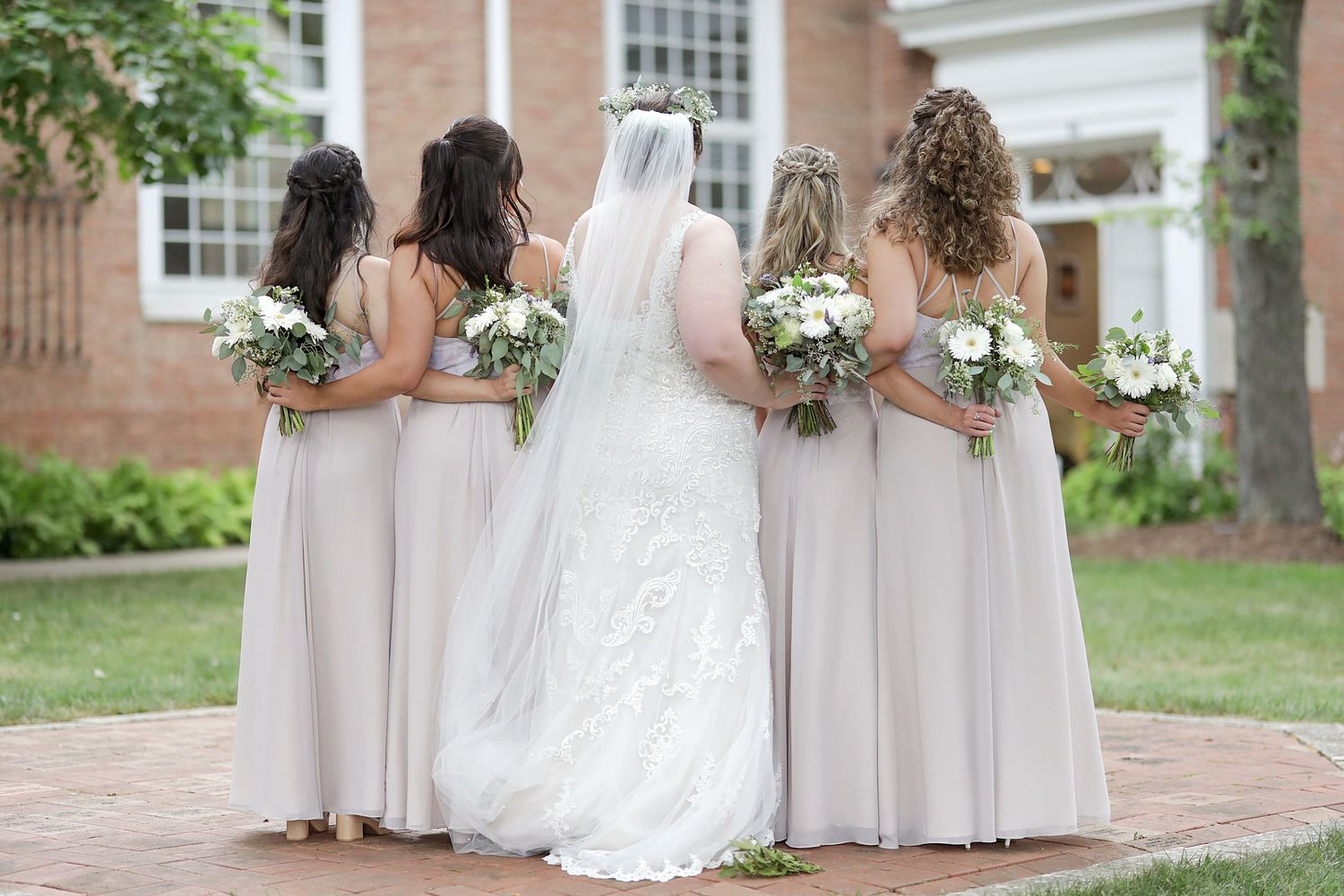 Wedding party in soft blush bridesmaid dresses and white bridal gown stand together holding floral bouquets on brick path.
