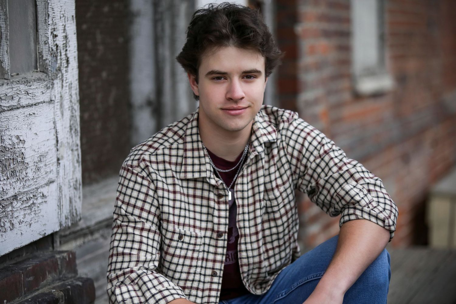 Young person in checkered shirt poses casually against rustic brick wall background in black and white photo series.