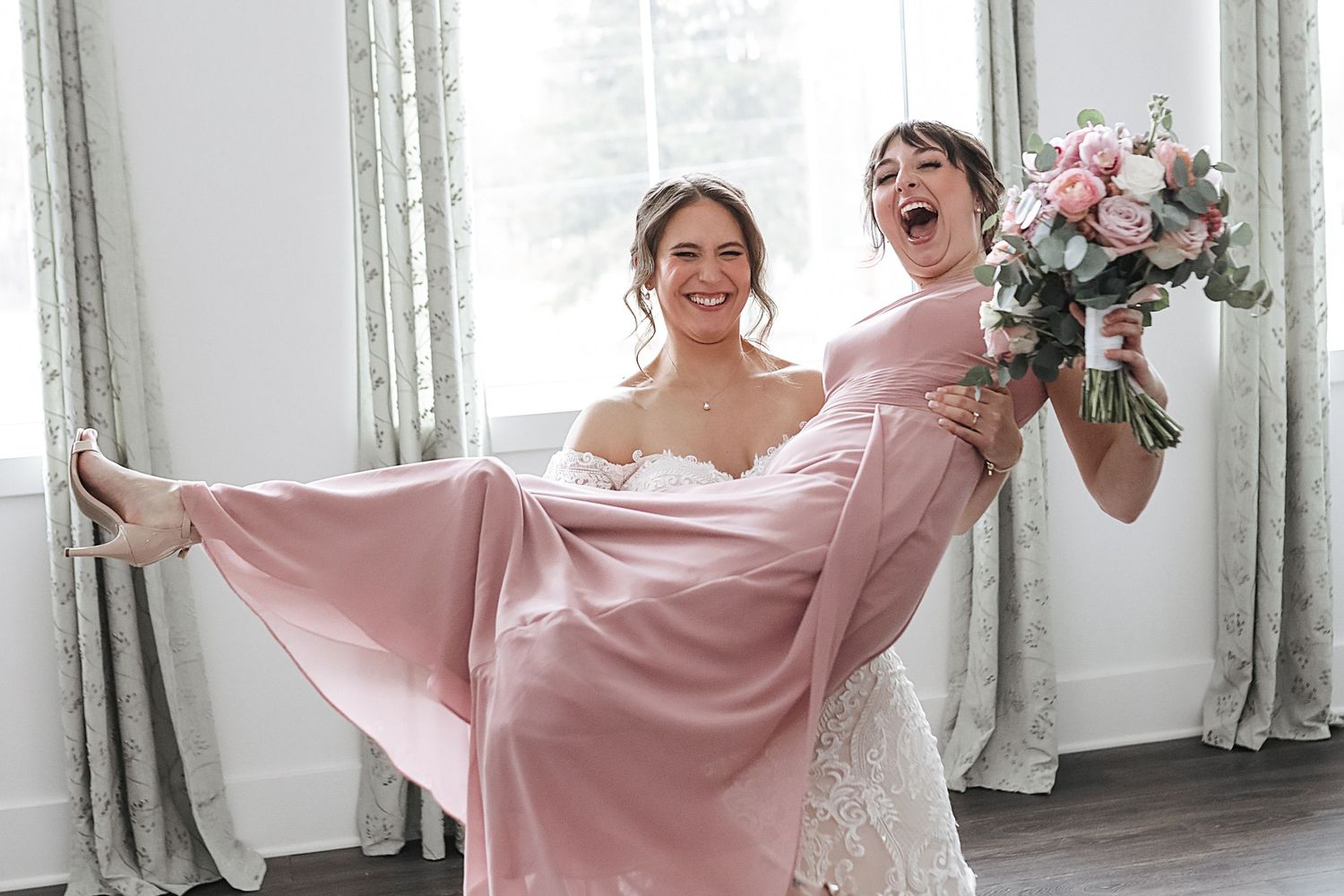 Bridesmaids in flowing pink dresses laugh and twirl together during a joyful wedding photo session.
