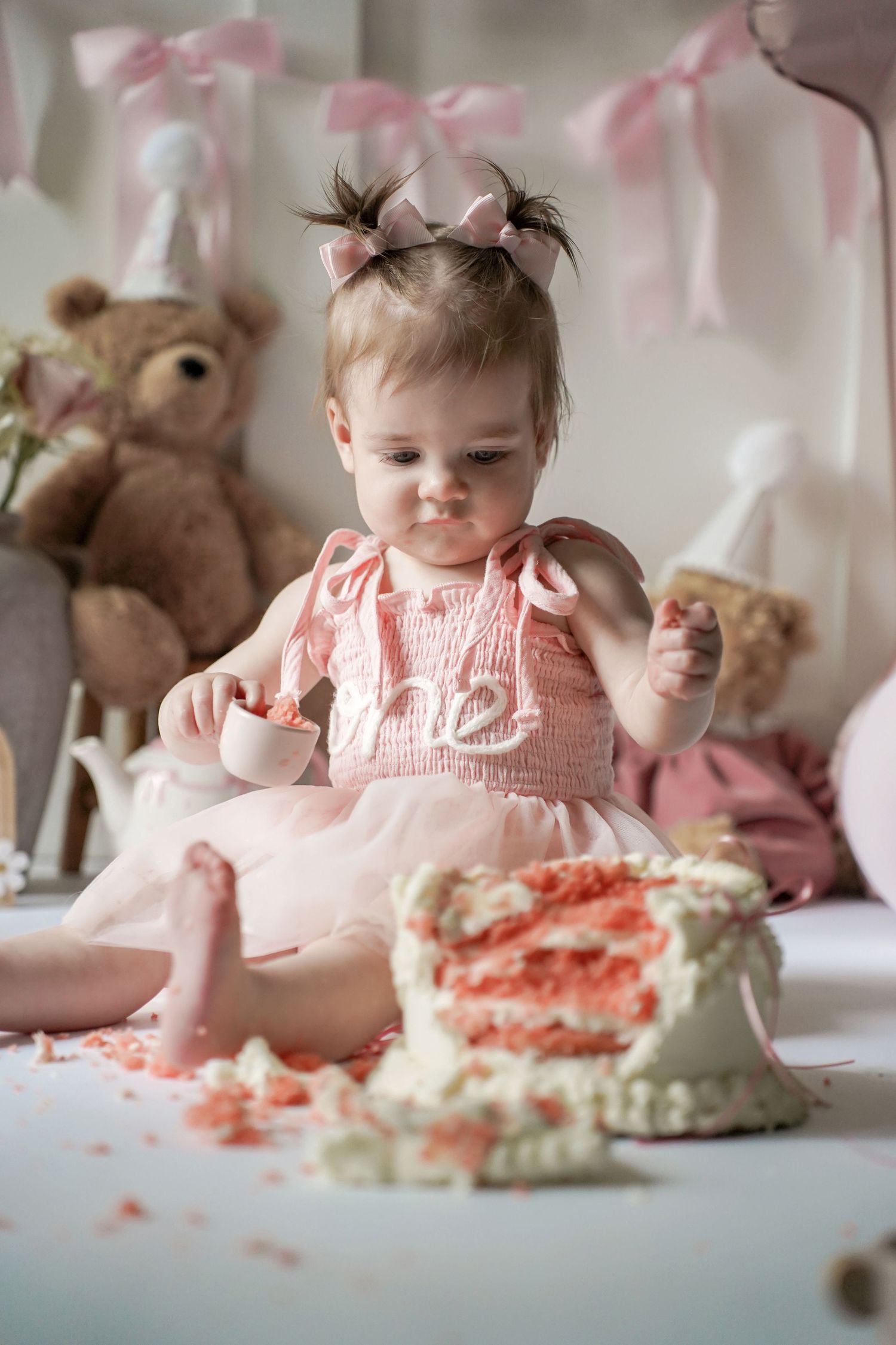 A pink tulle dress-clad baby sits among stuffed animals with a smashed birthday cake during a cake smash photo session.