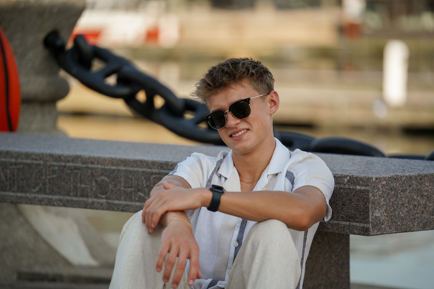 A person in sunglasses and a white polo shirt sits casually outdoors while leaning against a wall in summer sunlight.