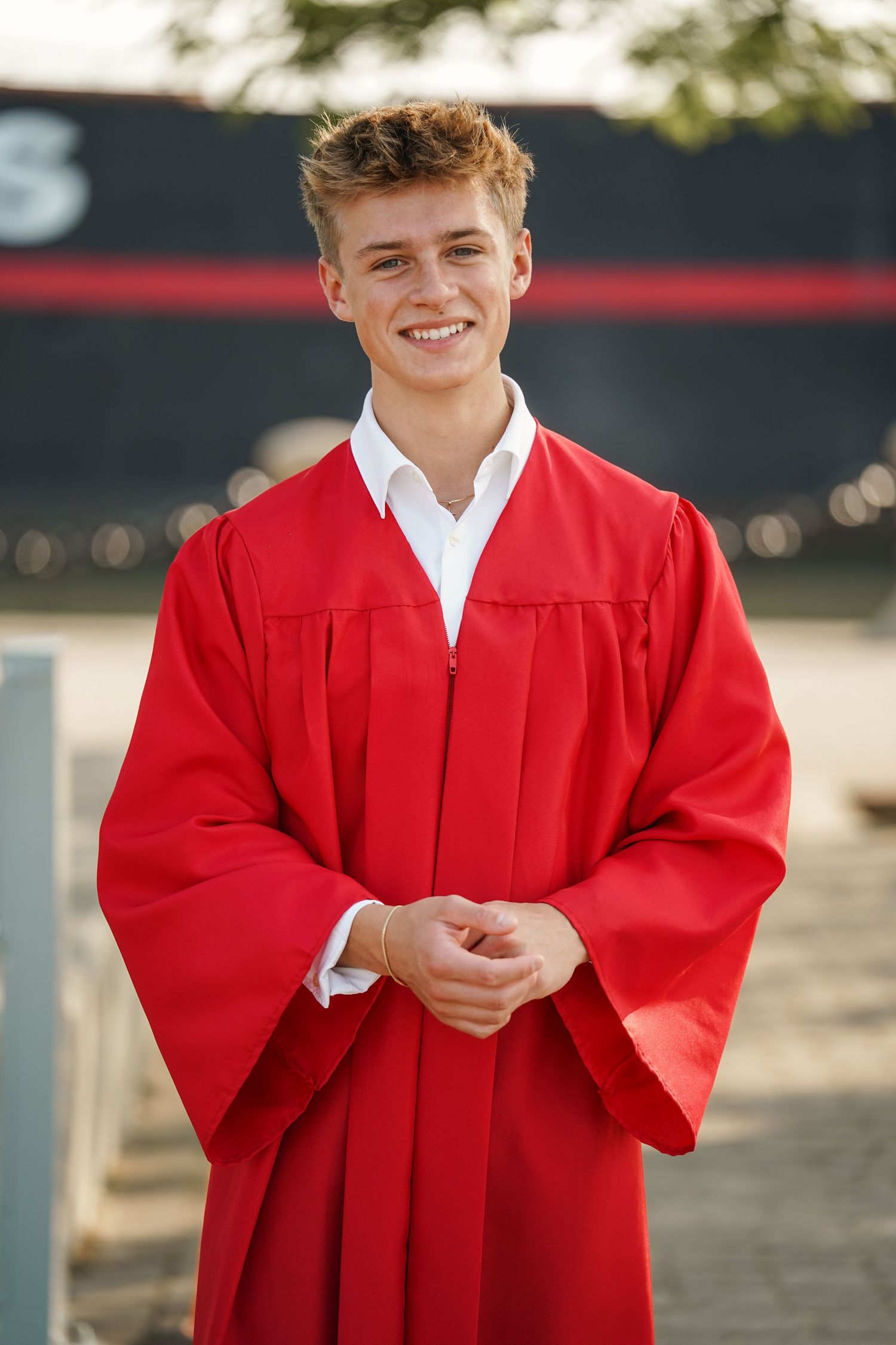 Graduate wearing bright red graduation gown and white collared shirt smiles while posing outdoors.