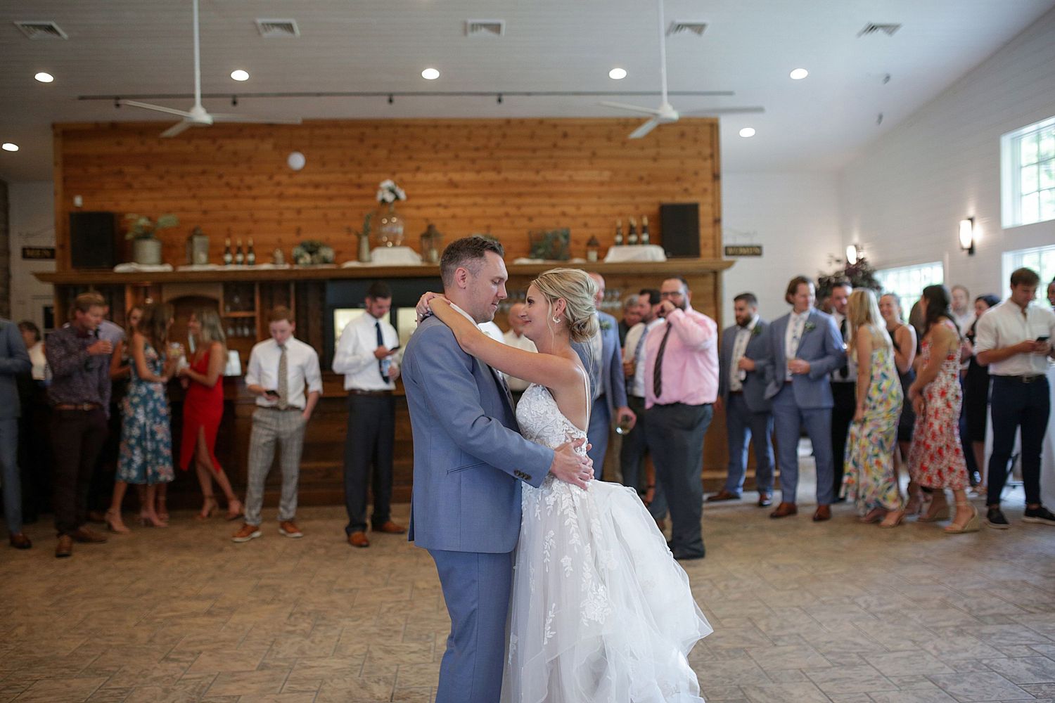 A couple shares their first dance in a rustic venue with wooden walls and guests looking on.