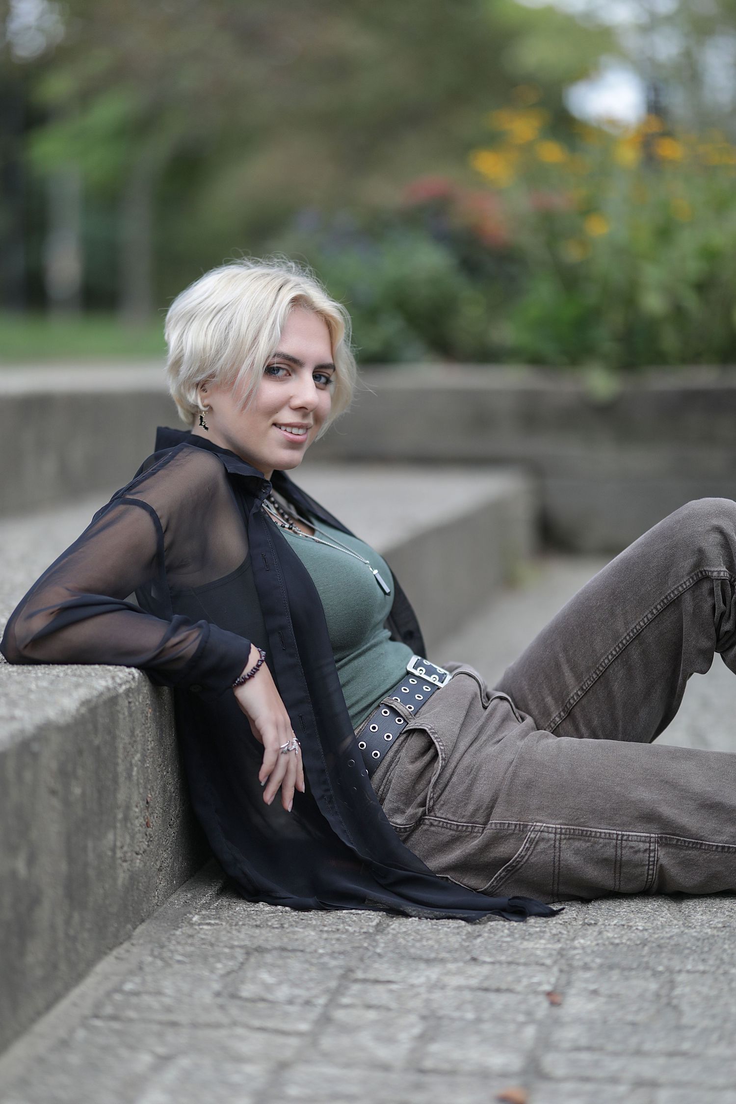 A person in sheer black top and cargo pants reclines casually on concrete steps outdoors with blurred greenery in background.