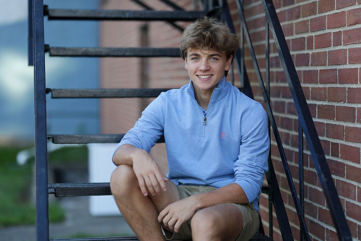 A person in a light blue quarter-zip sweater sits on outdoor stairs against a brick wall.