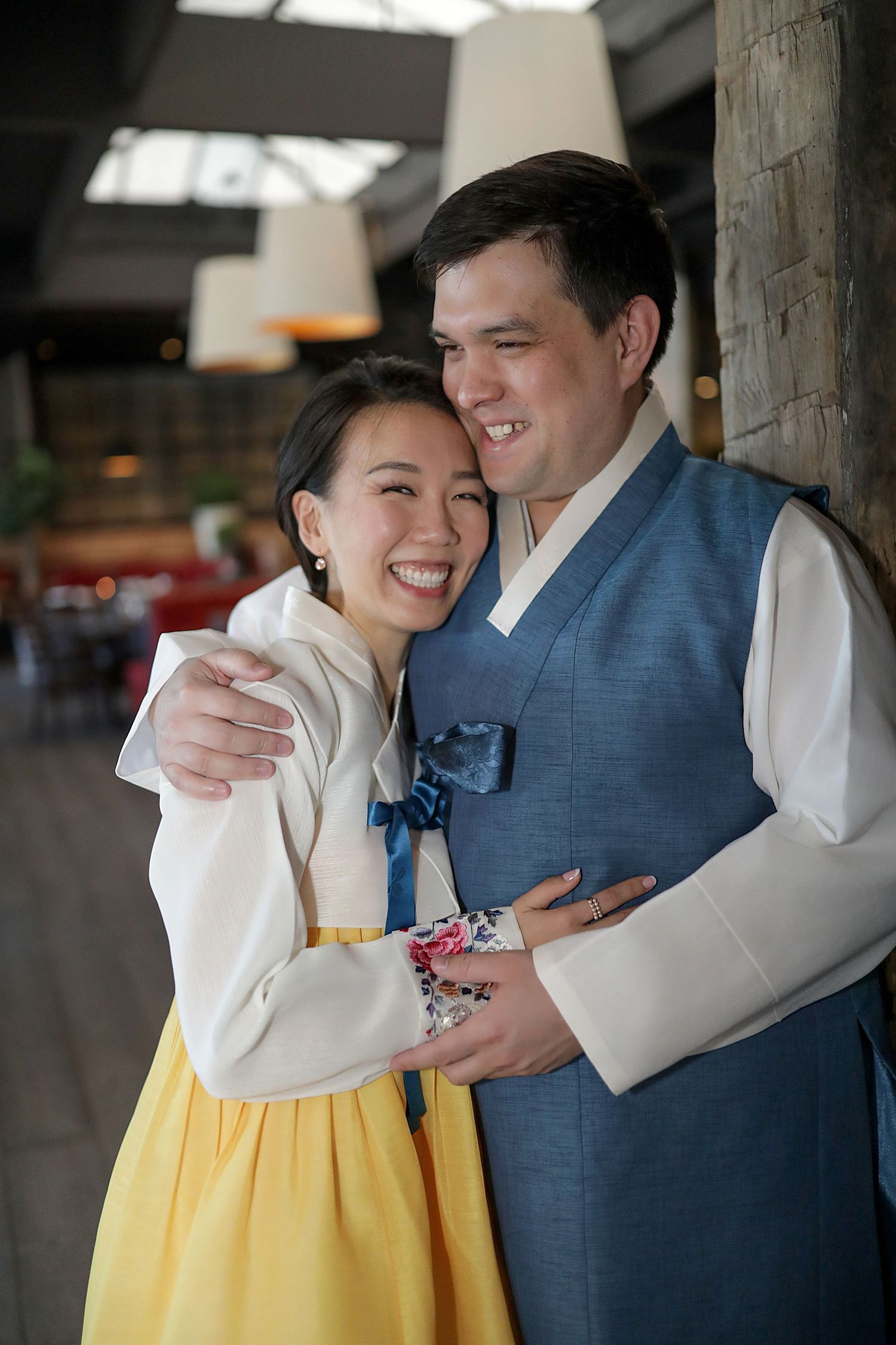 A couple embraces in traditional Korean hanbok attire, with the woman wearing white and yellow and the man in a blue vest.