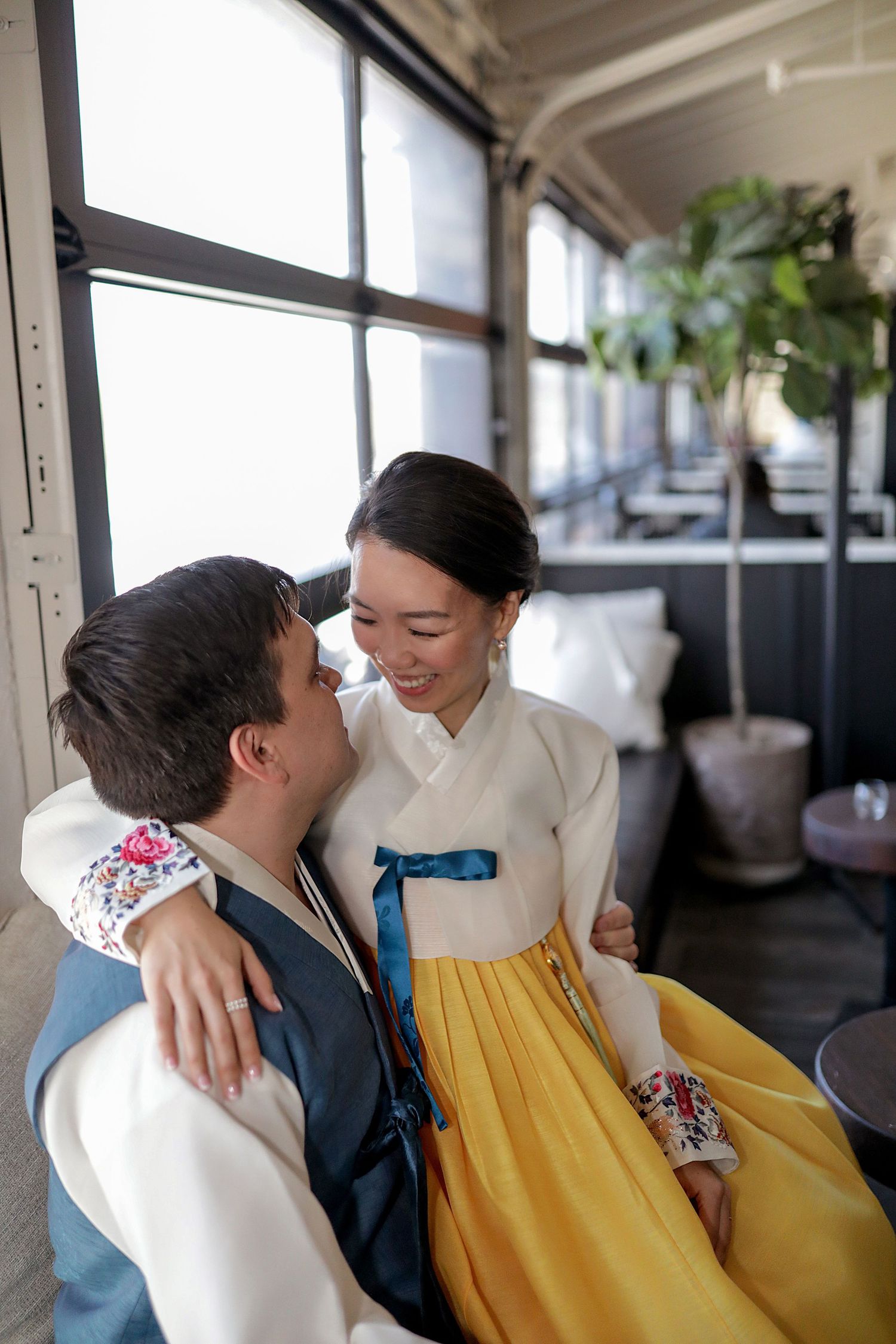 A couple in traditional Korean hanbok attire share an intimate moment by a window in natural lighting.
