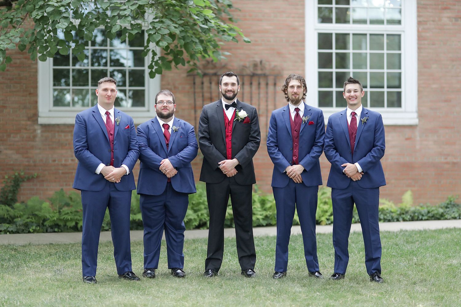 A group of groomsmen in navy suits and red ties pose together on grass in front of a brick building.