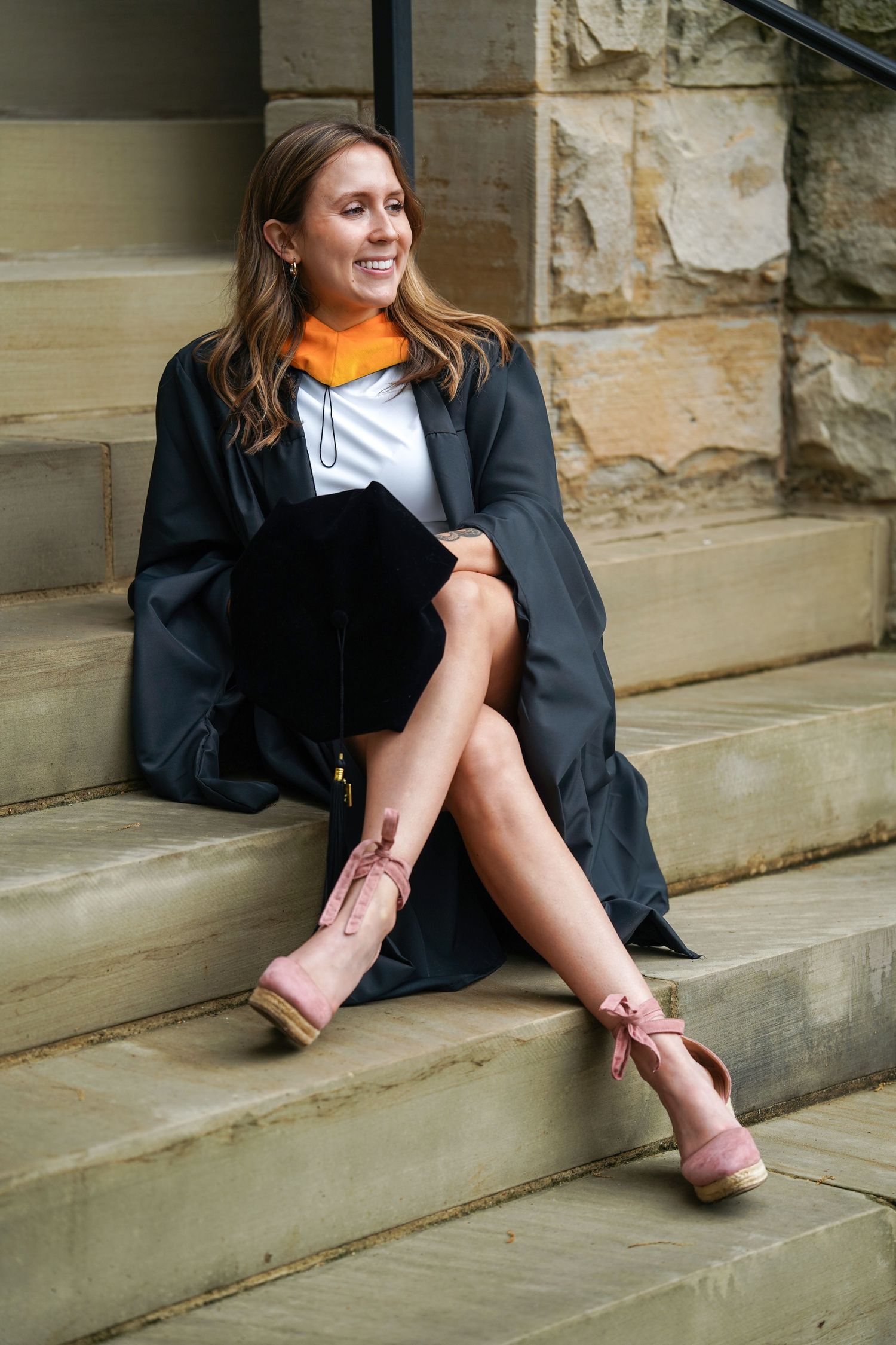 Recent graduate in cap and gown sitting on stone steps wearing pink strapped heels and smiling joyfully.