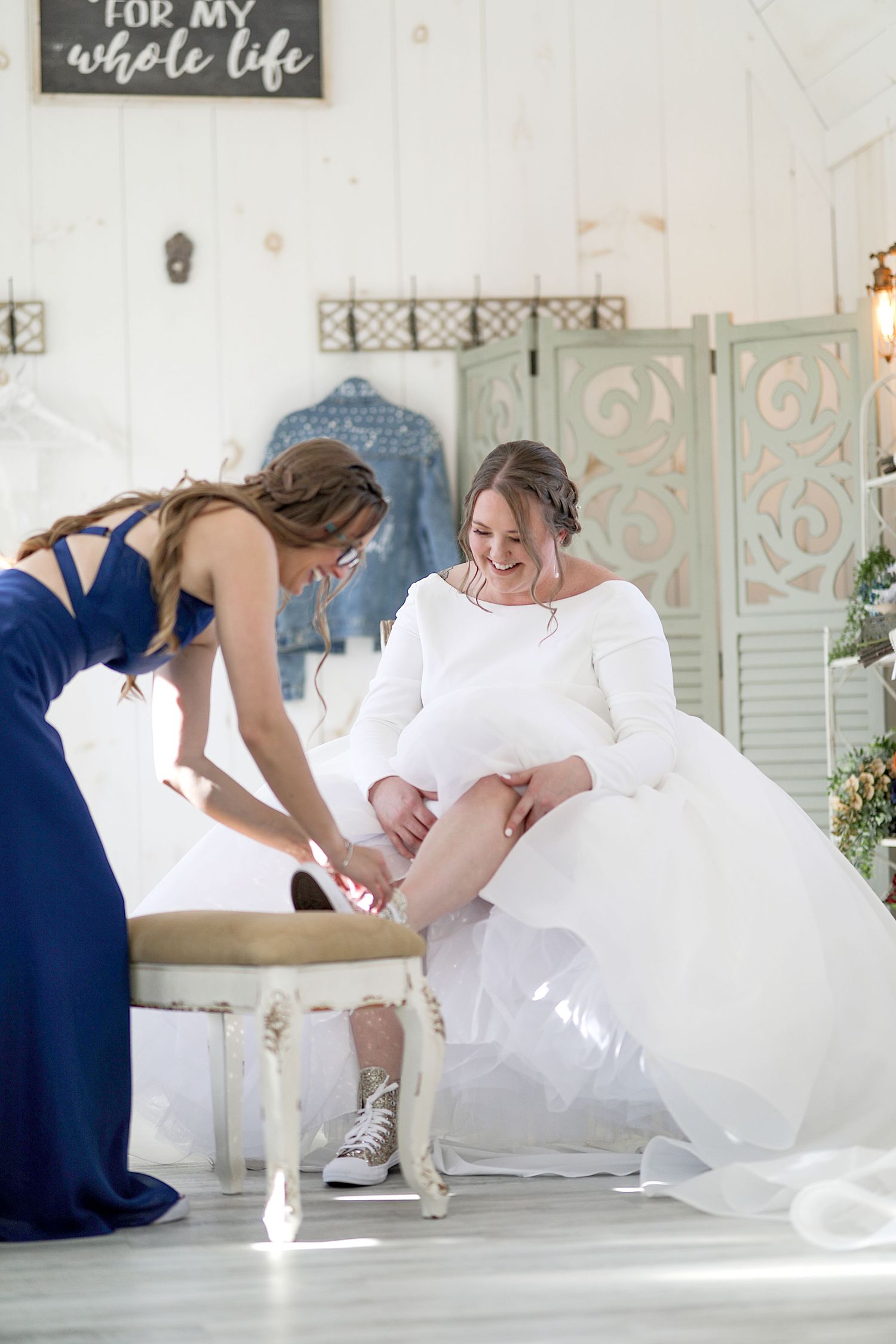A bride in a white dress with sneakers has her dress adjusted in a light-filled room with decorative screens.
