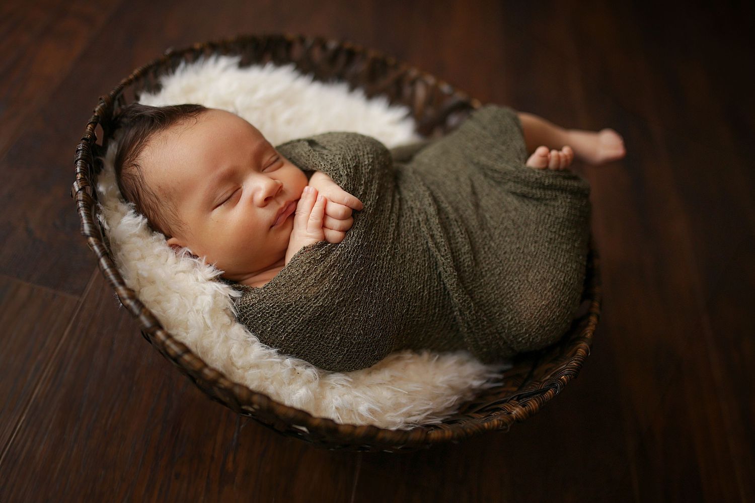 Sleeping newborn wrapped in olive green swaddle rests in cozy basket with furry lining. Newborn photographer Medina, Ohio.