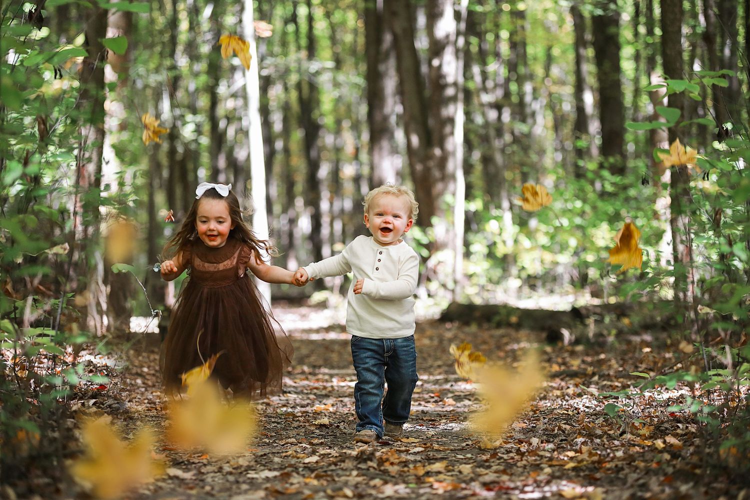 Two children running through autumn forest with golden leaves falling around them.  Family Photography in Medina Ohio.