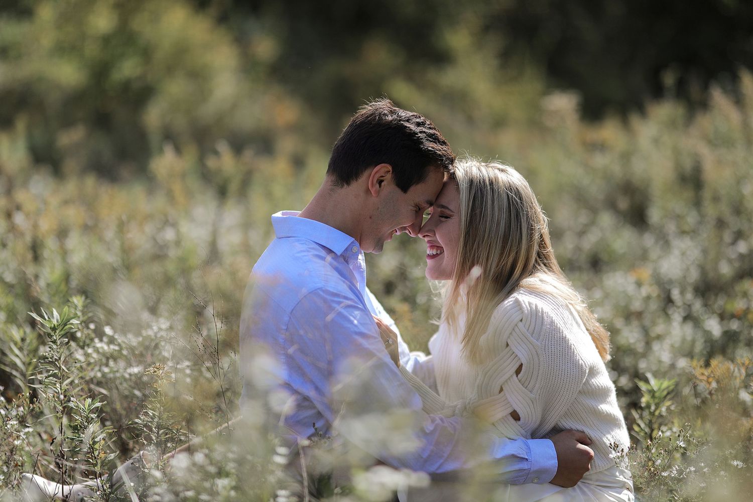 Romantic couple sharing an intimate moment in a sunlit wildflower field wearing white and blue clothing.