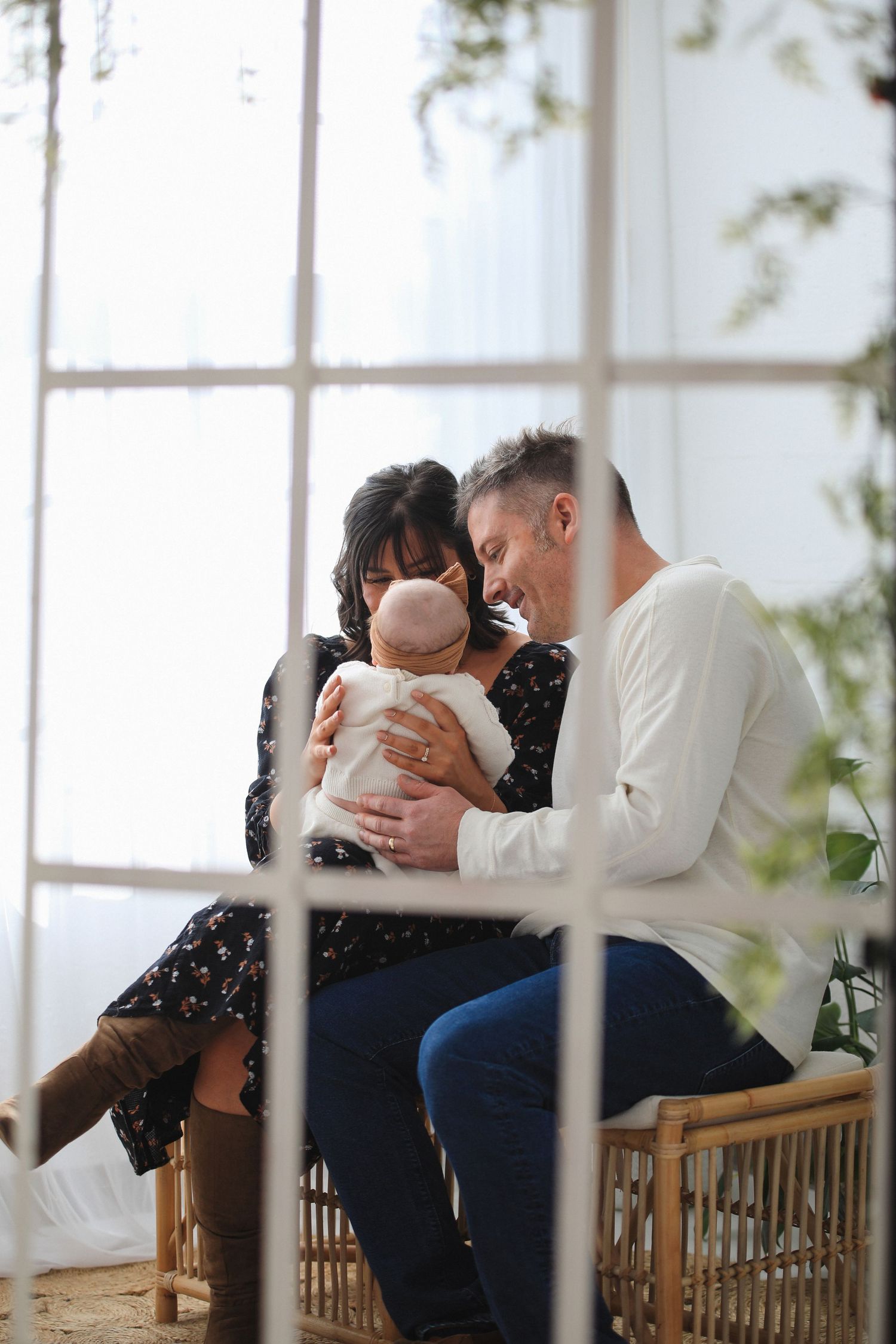 Family sits together on wooden bench in bright sunlit room with large windows and white walls.