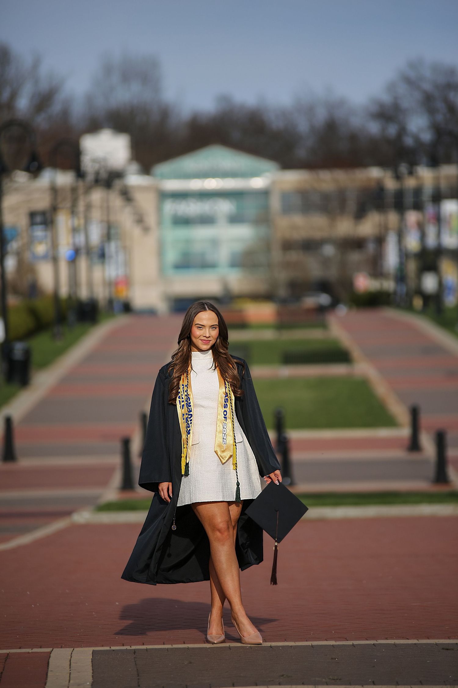 Graduate in cap and gown poses on campus walkway with decorative stole during outdoor graduation photo session.