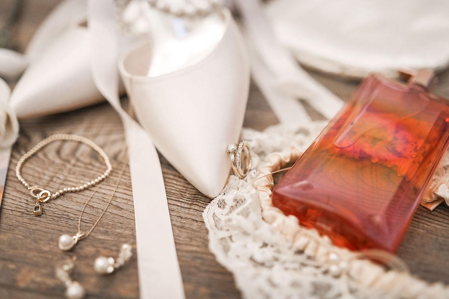 Elegant wedding details including white ribbon, lace, pearls, and pink perfume bottles arranged on rustic wooden surface.
