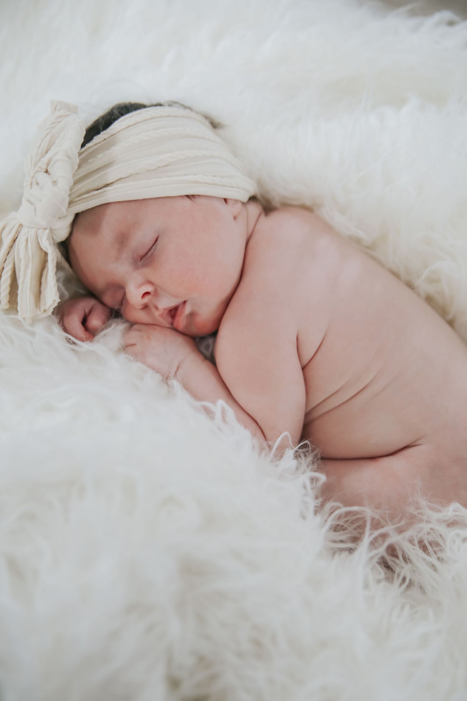 A peaceful newborn baby sleeps soundly on soft white fur blankets in a dreamy professional photography setting.