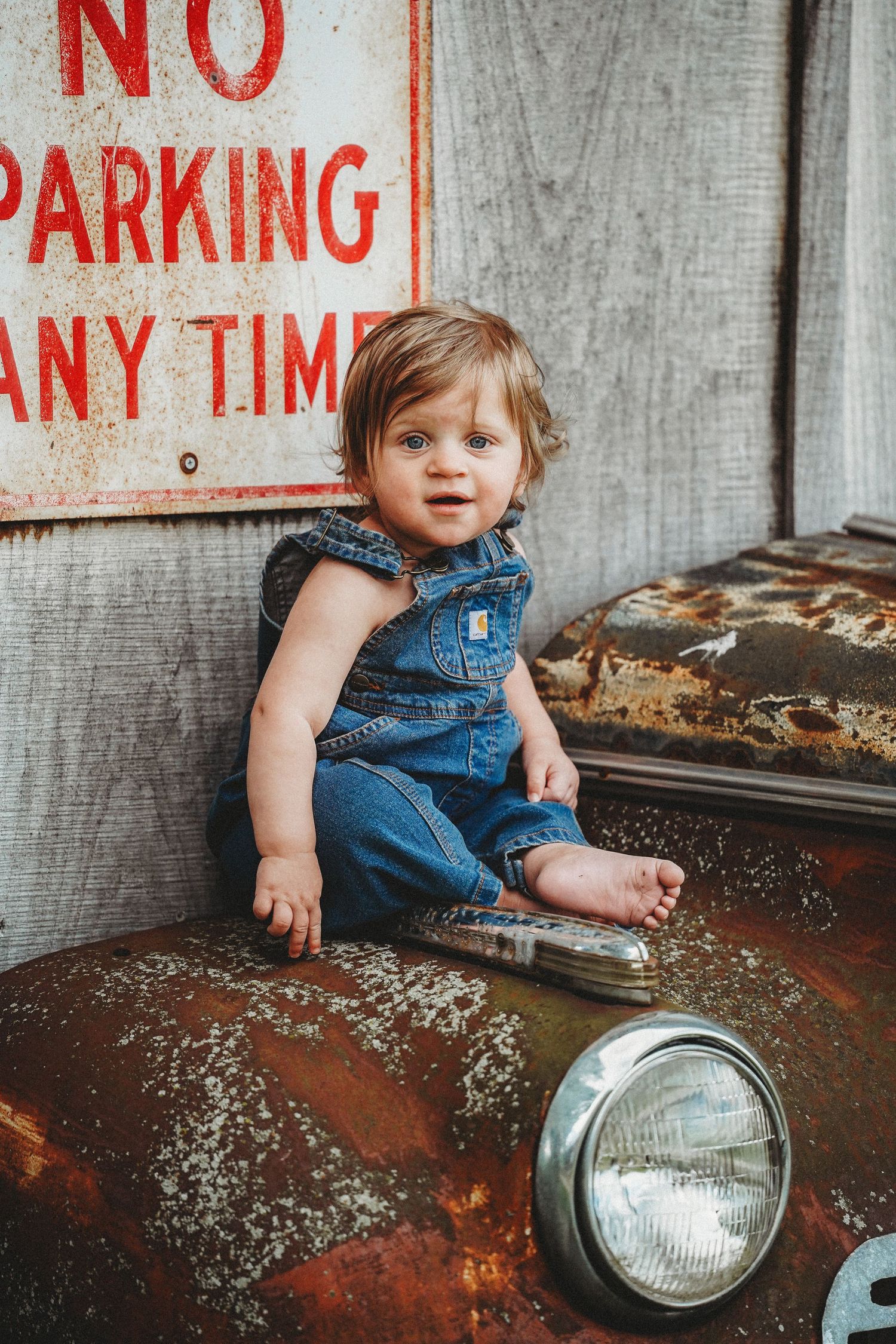 A child in denim overalls sits next to a vintage car headlight against a concrete wall with No Parking signs.
