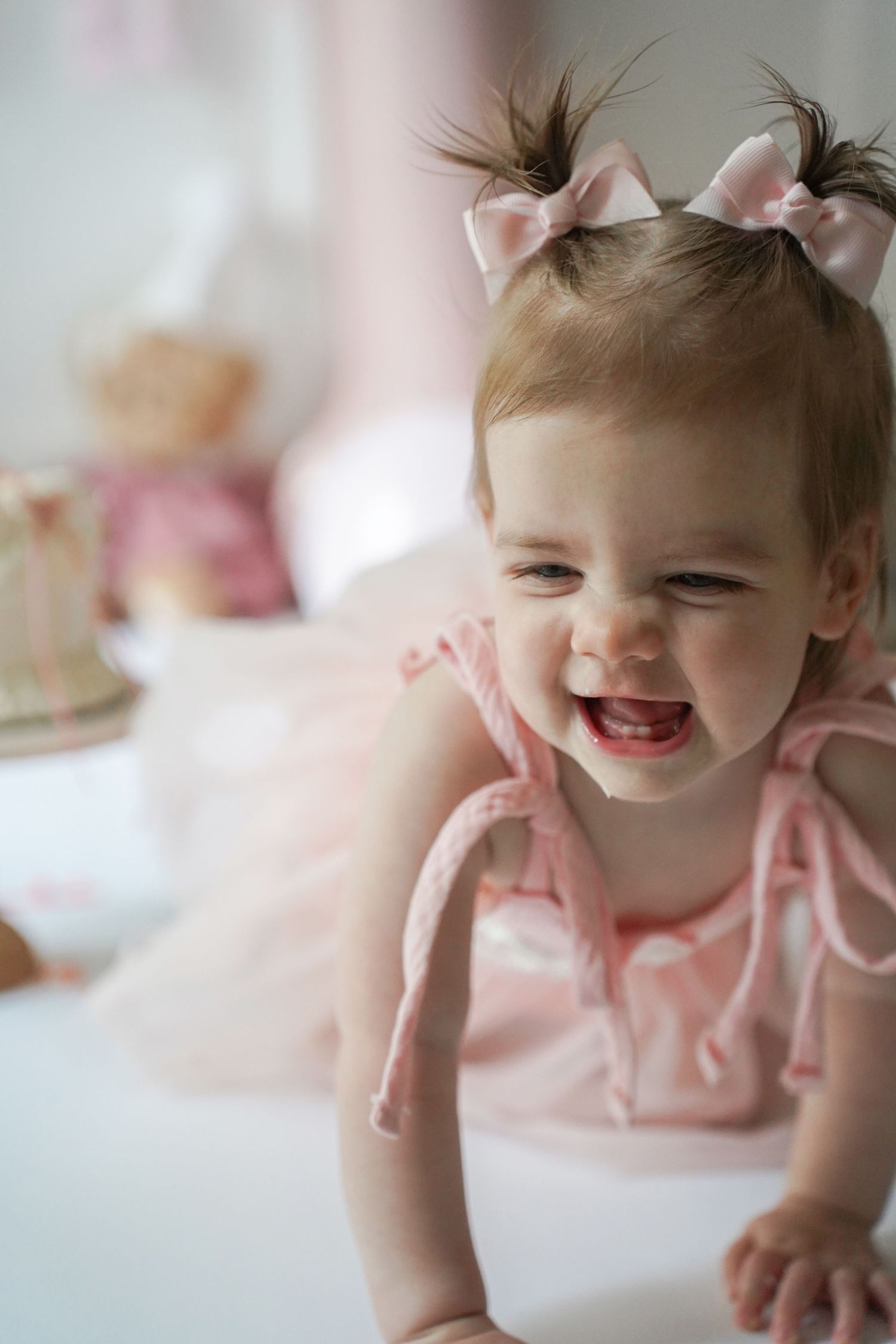 A young toddler wearing pink hair bows and a pink outfit smiles joyfully while crawling.