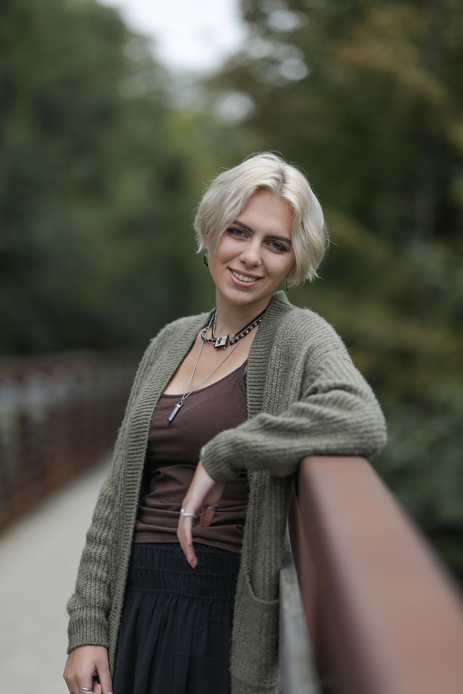 A person wearing an olive green cardigan and brown tank top poses outdoors with a blurred natural background.