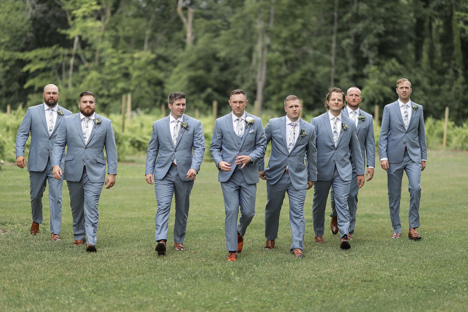 Groomsmen in matching light grey suits walk together on grassy lawn during outdoor wedding.