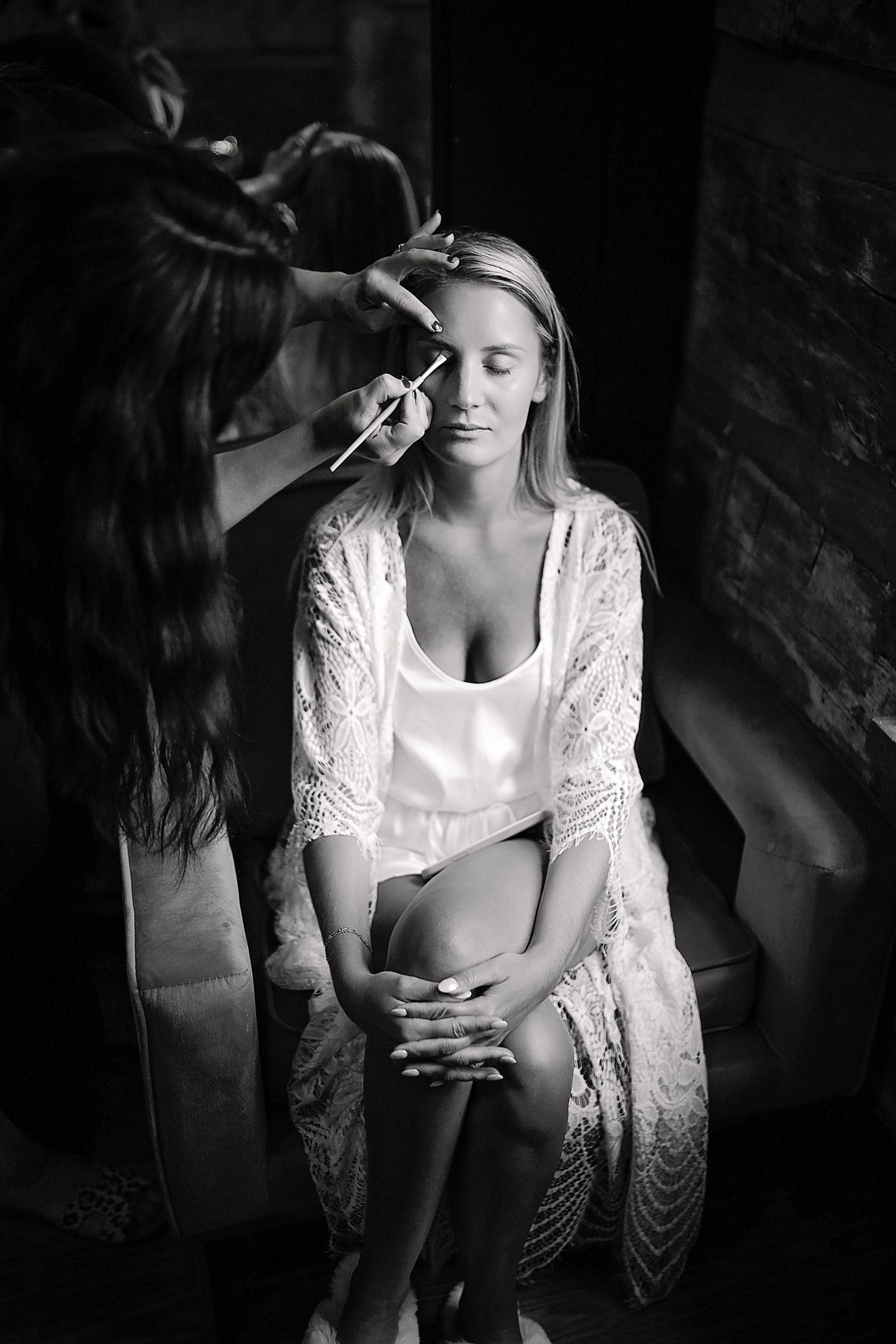 Elegant black and white photo of bridal preparations with makeup being applied in delicate lace robe.