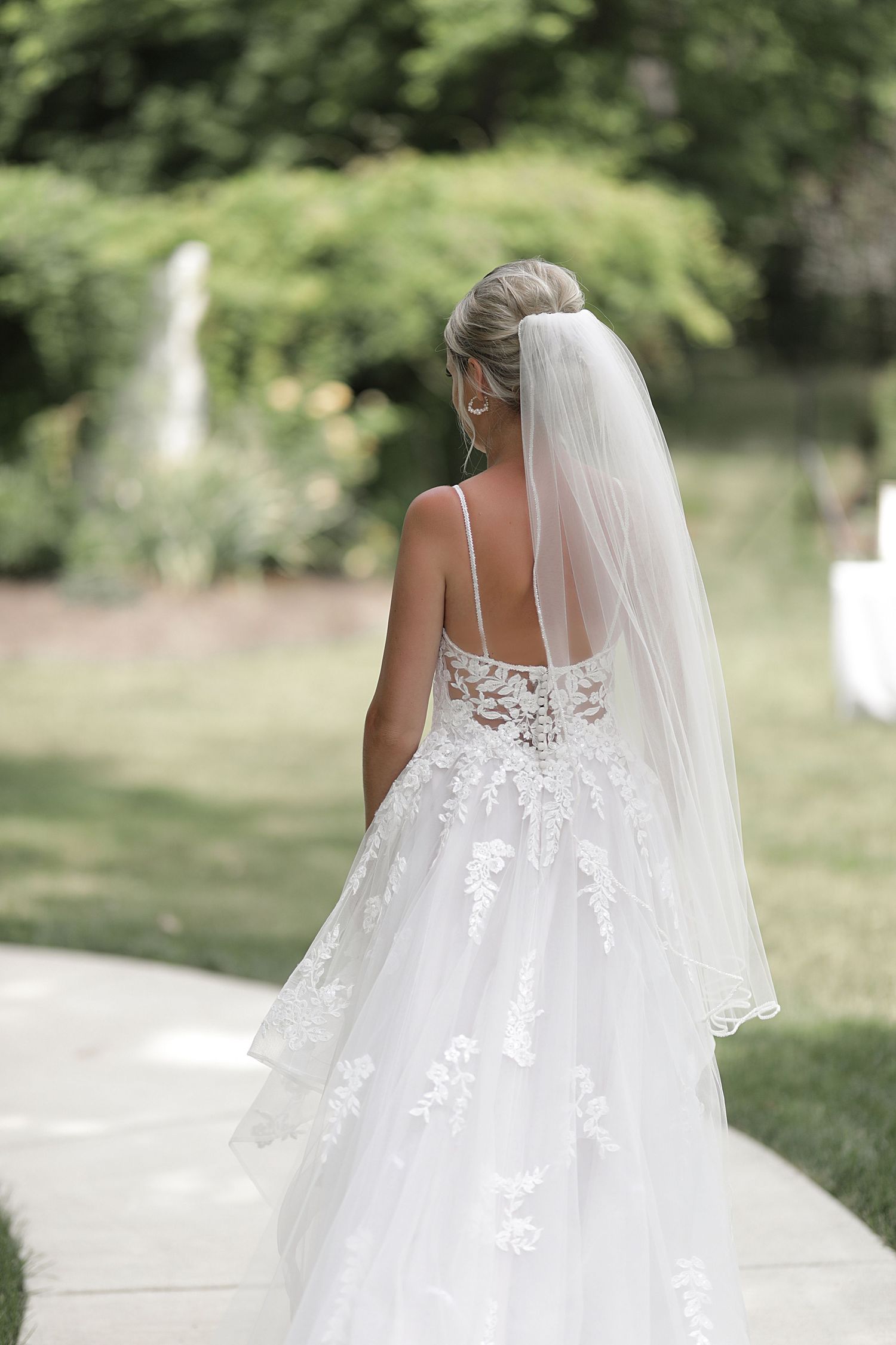 Beautiful bride in a lace wedding dress with flowing veil poses outdoors in a garden setting.