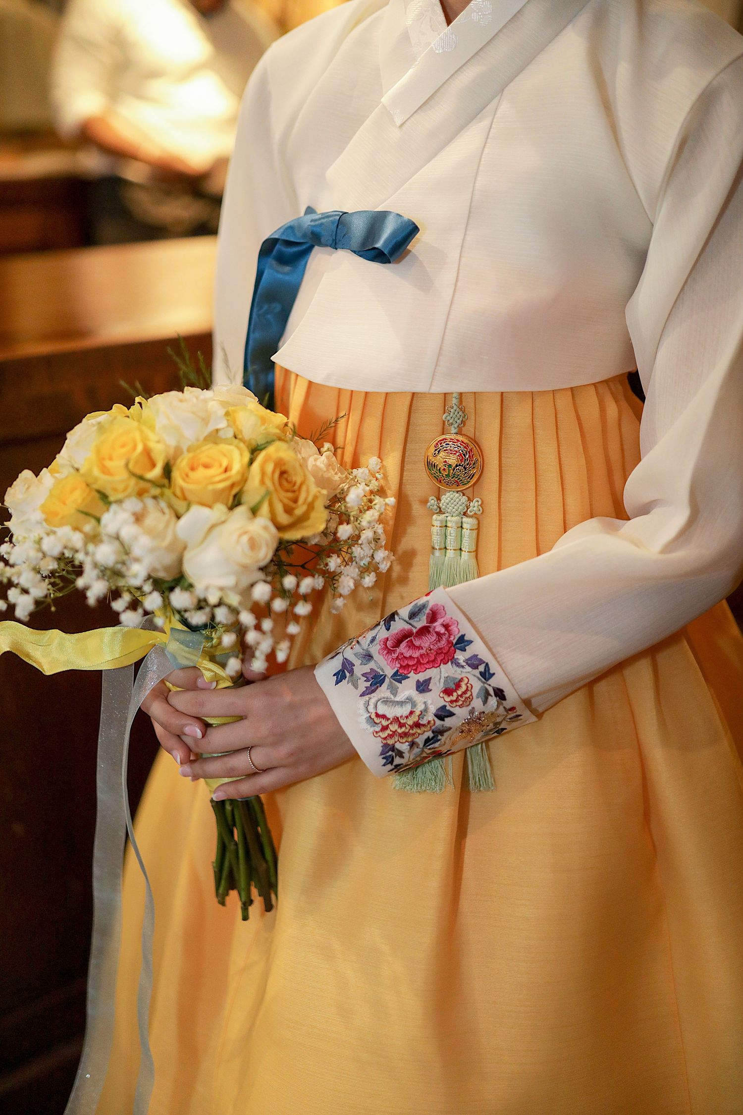 Traditional Korean hanbok outfit in white and yellow with decorative corsage and bouquet of yellow roses and white flowers.