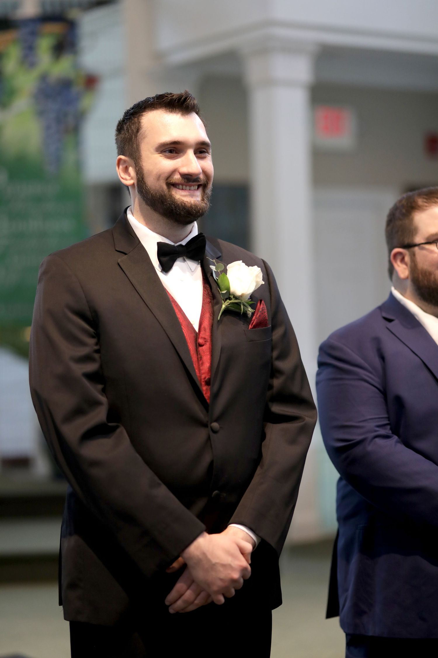 Groomsmen wearing formal suits stand in line during a wedding ceremony with black tuxedos and red accents.