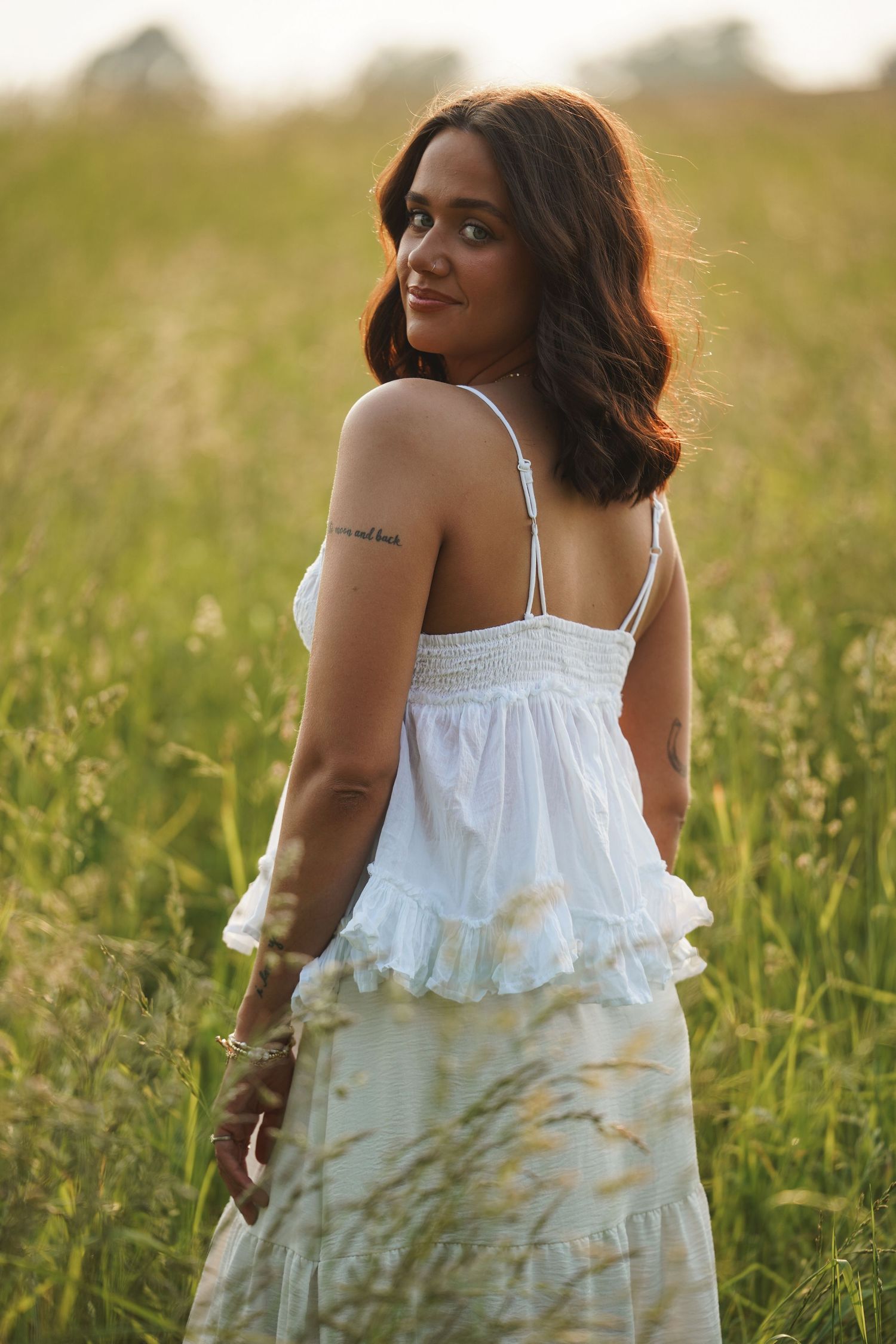 A person in a flowing white summer dress poses in a sunlit meadow during golden hour.