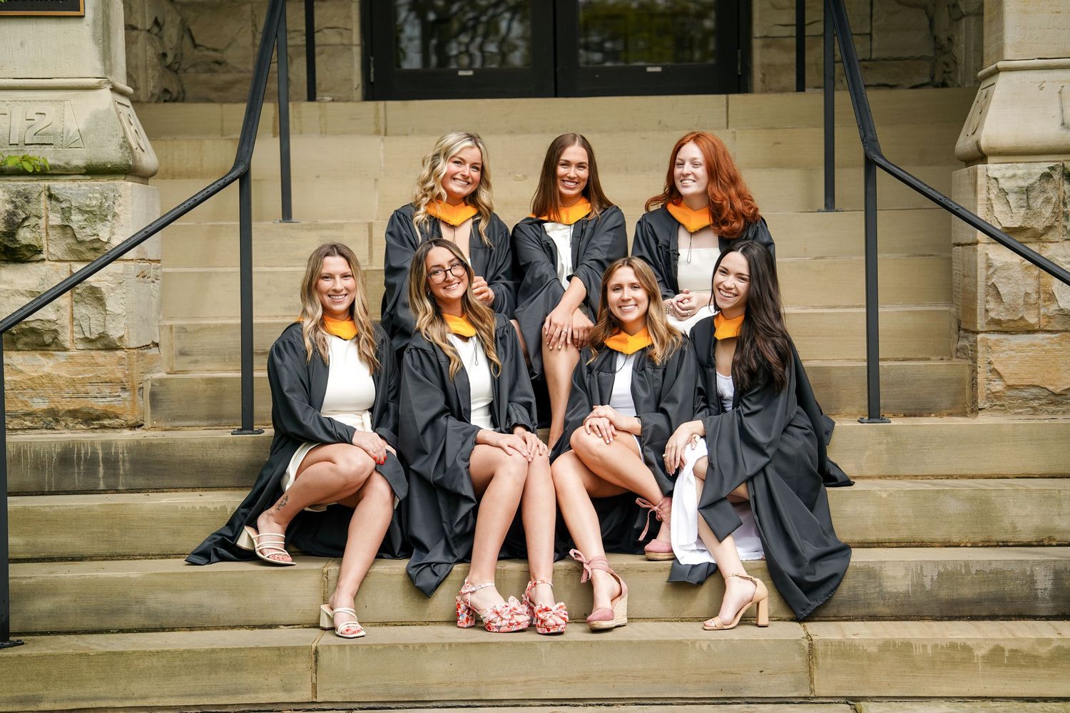 A group of graduates in black robes sit together on stone steps of a building in celebratory poses.
