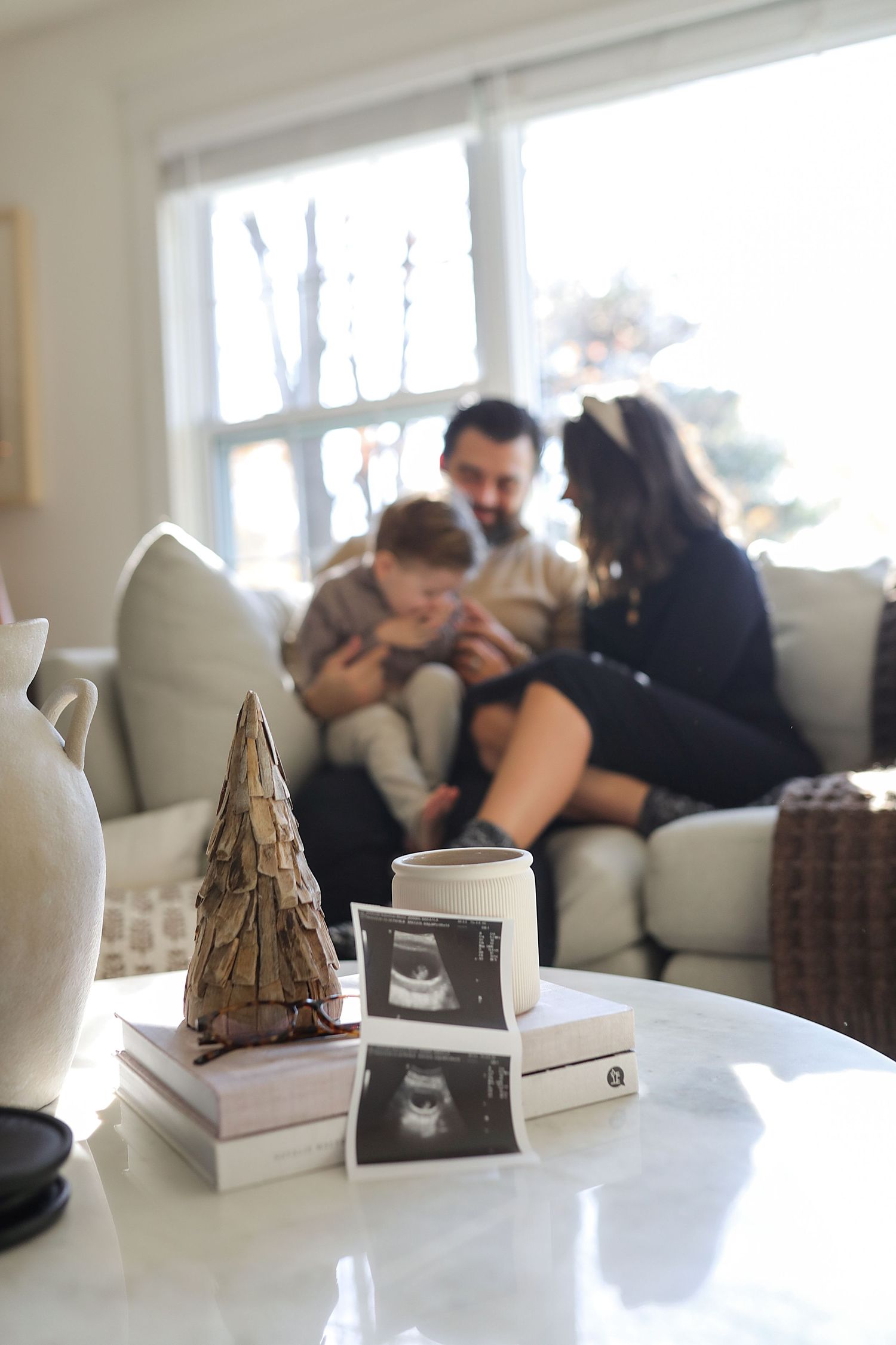 In a bright living room, a family sits together on a couch while ultrasound photos and a decorative tree rest on a table.