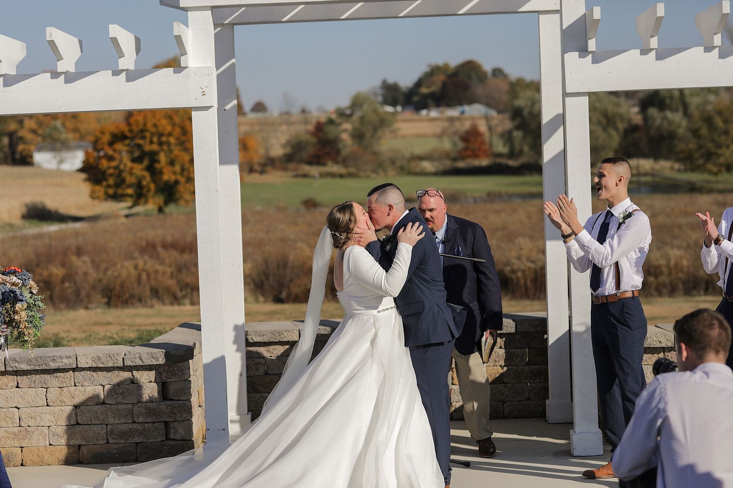 Wedding ceremony setup with white pergola and stone wall backdrop in fall landscape.