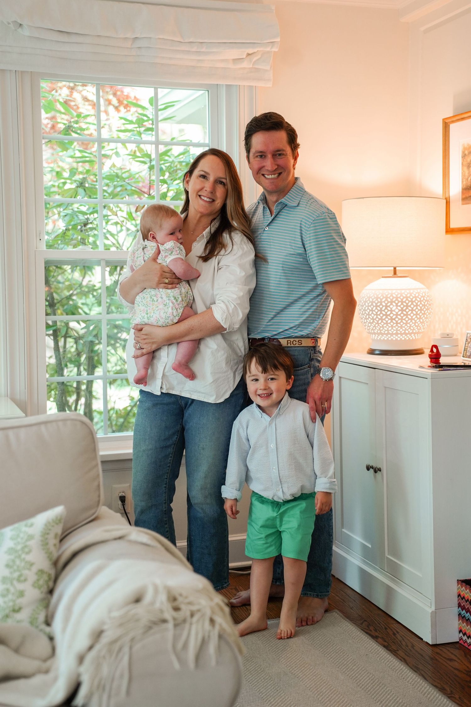 A family stands together by a window in a cozy living room with white cabinets and warm lighting.